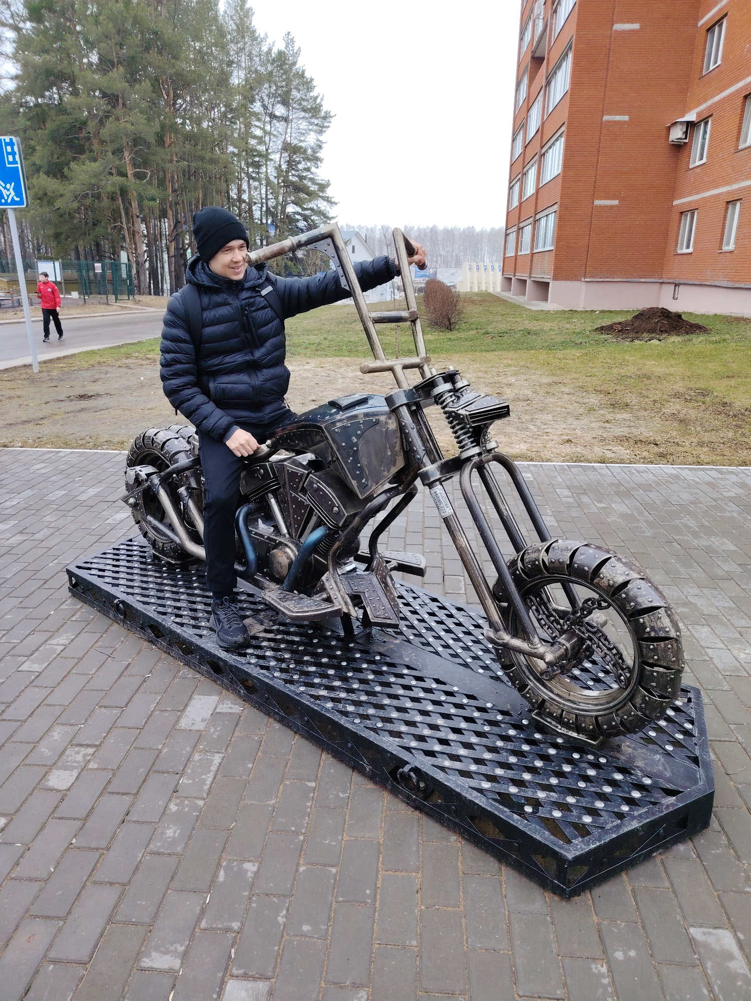 A young man is posing on a metal motorcycle sculpture in Chaikovsky, Russia. He is wearing a black puffer jacket and a black beanie. He is sitting on the motorcycle's seat with one foot on the footrest and the other on the metal platform beneath. The motorcycle sculpture is made of dark metal with exposed engine components and fabricated wheels that give it a rugged look. It is mounted on a black metal platform with a diamond-patterned grate surface.
The scene is set outdoors on a paved area, likely a pedestrian walkway. Behind the man, there is a background of evergreen trees and a multi-story brick building with windows. A man in a red jacket is seen walking in the far background near a sign with blue symbols. The sky appears overcast, indicating a potentially cool or mild day. The overall setting seems to be a public space, possibly a park or a town square. Patches of dormant grass are visible behind the motorcycle sculpture.