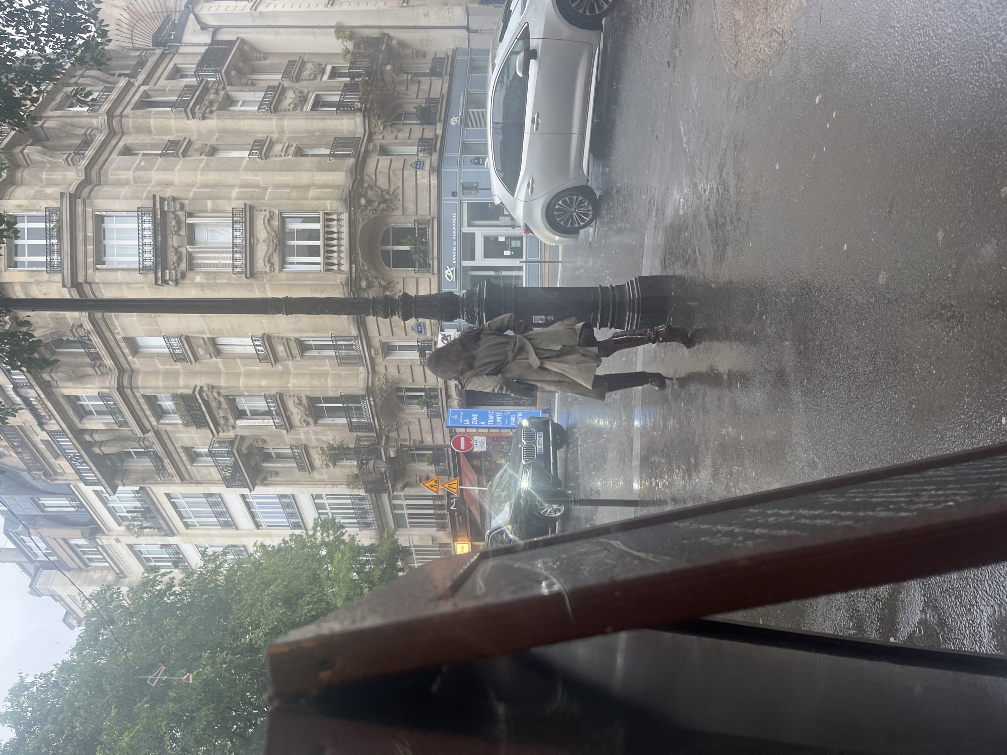 The image depicts a street scene in Paris, France during a rainy day. A person wearing a light-colored trench coat and dark pants is running across the wet sidewalk toward the left side of the frame, seemingly attempting to cross the street. A black vintage lamp post stands between the person and the camera.
Several cars, including a silver hatchback and a black sedan, are parked along the right side of the street. Traffic signs, including one indicating a limited traffic zone in Paris, are visible near the black sedan. Buildings with traditional Parisian architecture, featuring ornate window details and balconies, line the street. The Banque et Assurances is visible behind the silver hatchback.
The photograph is taken from inside what appears to be a restaurant or café, with a part of a table or menu visible in the foreground. The rain obscures some details, giving the scene a slightly blurred, atmospheric quality.