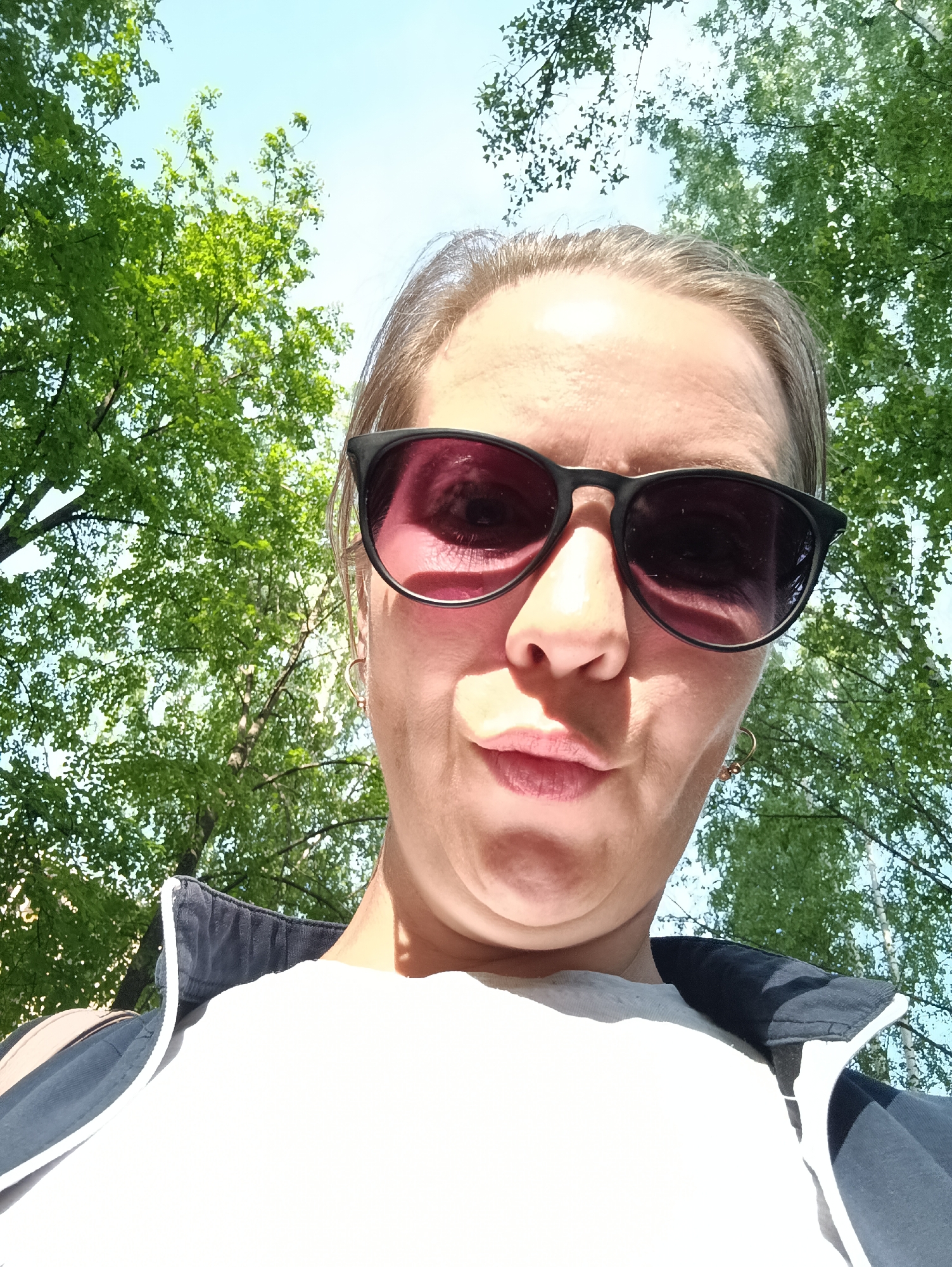 A selfie taken in an outdoor environment, likely a park or wooded area based on the presence of trees with green leaves against a bright sky, possibly taken in the city of Chaikovsky, Russia. The focal point is a woman wearing dark sunglasses, a white t-shirt, and a dark jacket. Her expression appears to be a slight pout or duck-like facial gesture.