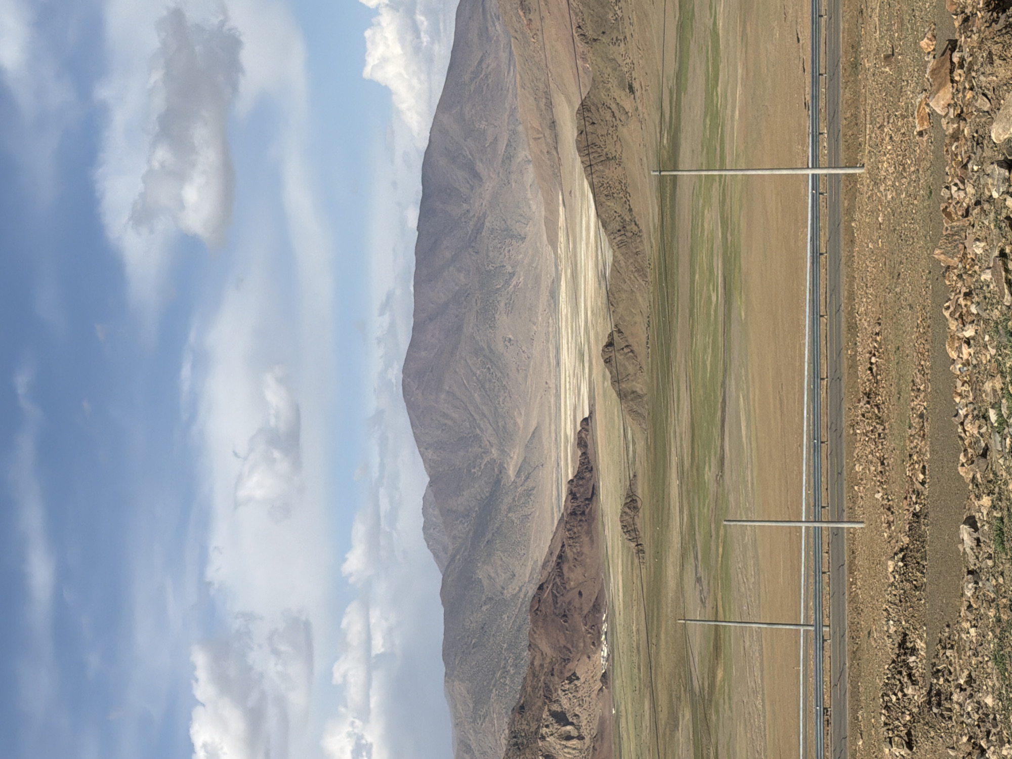 The image depicts a landscape in Gyirong County, China, featuring a tall, rugged mountain range on the left. A broad, dry basin lies at the base of the mountains. A road runs parallel to the mountains on the right, bordered by rocky terrain. Utility poles are spaced along the roadside. The sky is filled with clouds.