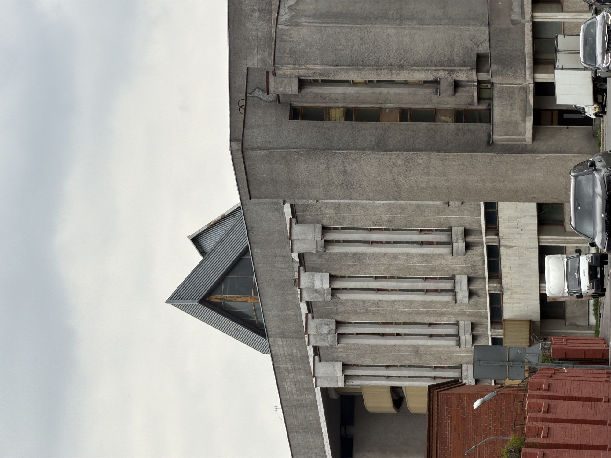The image depicts an architectural scene in Saint Petersburg, Russia, featuring a modern, brutalist-style building constructed primarily of gray concrete. The building has a distinct angular design with horizontal protrusions and visible window openings. A separate section of the building has a triangular apex with a glass window. To the side of the concrete structure is a brick building, and a street complete with street signs and a street lamp. Cars are parked along the street. The sky above is overcast, indicating cloudy weather conditions.