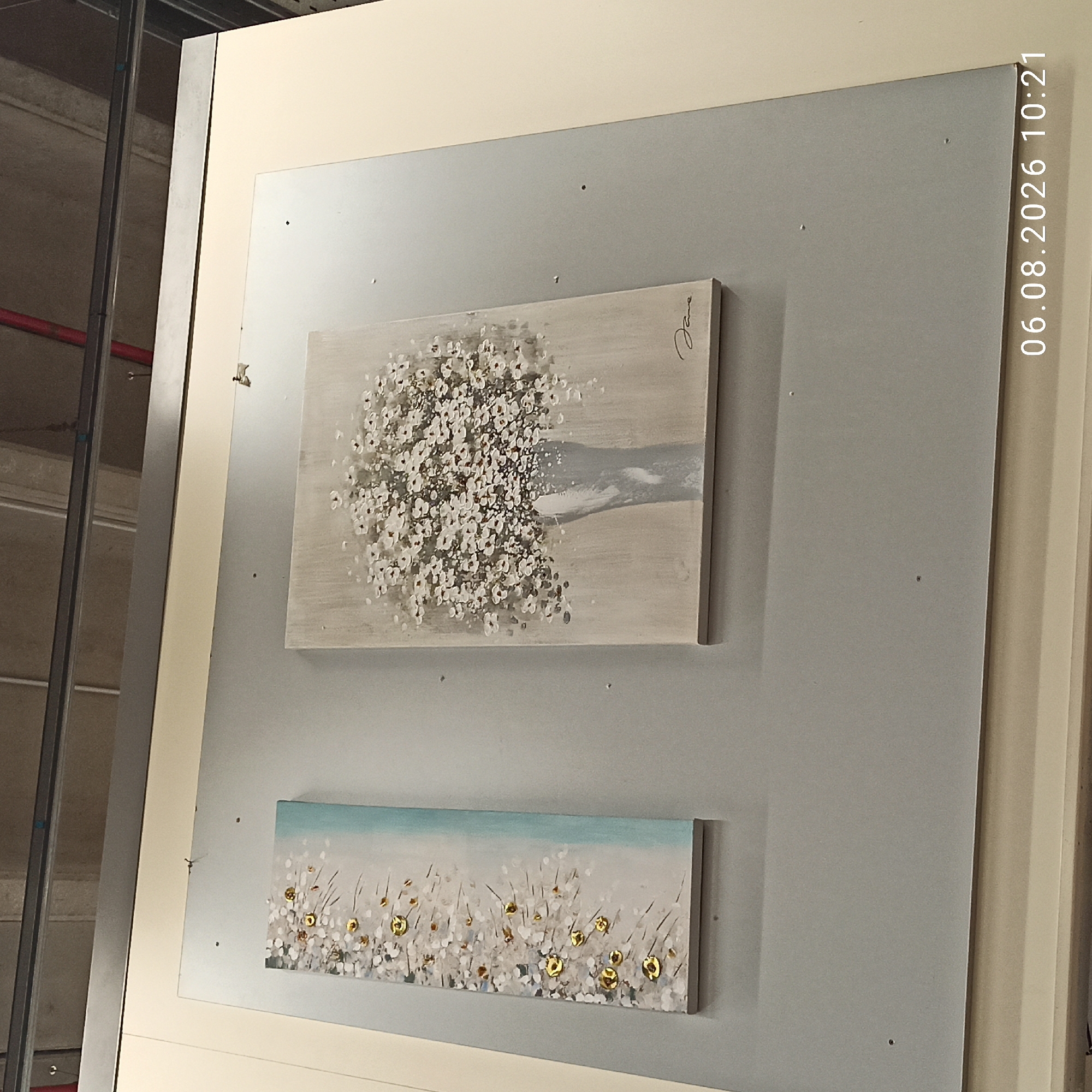 The image shows two framed paintings hung on a light grey wall. The painting on the left is a narrow, vertical piece with a white and grey floral design that transitions to a light blue background on the right. It features gold circular accents scattered throughout the floral pattern. The painting on the right is a larger, rectangular piece depicting a vase filled with numerous small white flowers against a neutral, textured background. The signature "Jane" is visible in the bottom right corner of this painting. Above the paintings, a metal beam runs horizontally across the frame, with exposed ceiling elements visible behind it. The bottom of the image displays a timestamp: "06.08.2026 10:21". The scene appears to be an indoor setting, possibly a gallery or a home, and there is no specific indication of its location in Bari, Italy, or any people present in the image.
