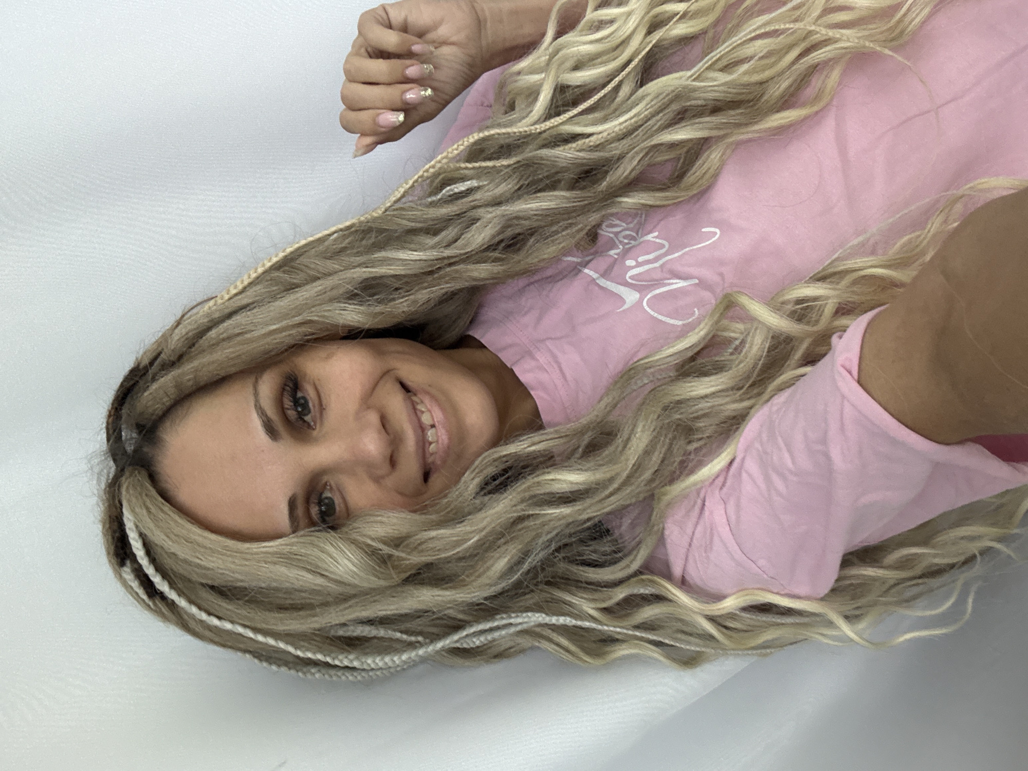 A smiling woman with long, wavy blonde hair mixed with white braids lies on a white surface. She is wearing a light pink shirt. The woman's hand is partially visible near the top of the frame, showing fingernails with a gold accent design. The image is a close-up shot with the woman's face and hair as the main focus.
Rusanovo, Russia is not discernible from the image.