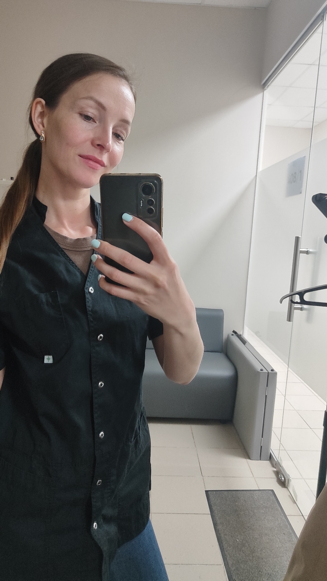 A woman is taking a selfie indoors, possibly in a medical facility or office in Chaikovsky, Russia. She is fair-skinned with brown hair pulled back in a ponytail and is wearing a dark green uniform-style shirt buttoned up over a neutral-toned undershirt. Her nails are painted a light blue color. She is holding a dark-colored smartphone with visible camera lenses. Behind her, there is a gray couch or bench and a glass-enclosed area with a door and a coat hanger. The flooring is light-colored tile with a gray mat near the door. There is lettering on the glass of the enclosed area, appearing to read "08/1".