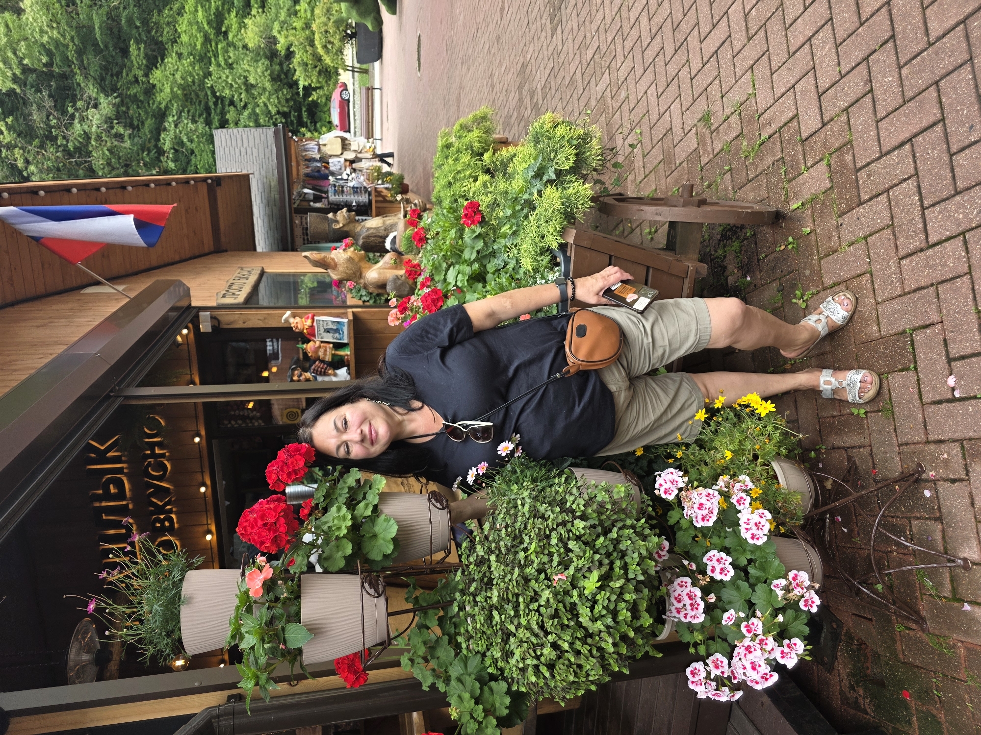 A woman of Middle Eastern descent, wearing a black t-shirt, khaki shorts, and silver sandals, stands in front of a store in Estosadok, Russia. She has dark hair and is smiling at the camera. She is holding a smartphone in her right hand and has a brown fanny pack around her waist. The store, adorned with a Russian flag, appears to sell souvenirs. Potted plants, including red and pink flowers, surround the woman. The ground is paved with reddish bricks. In the background, a red car and lush green trees are visible.
