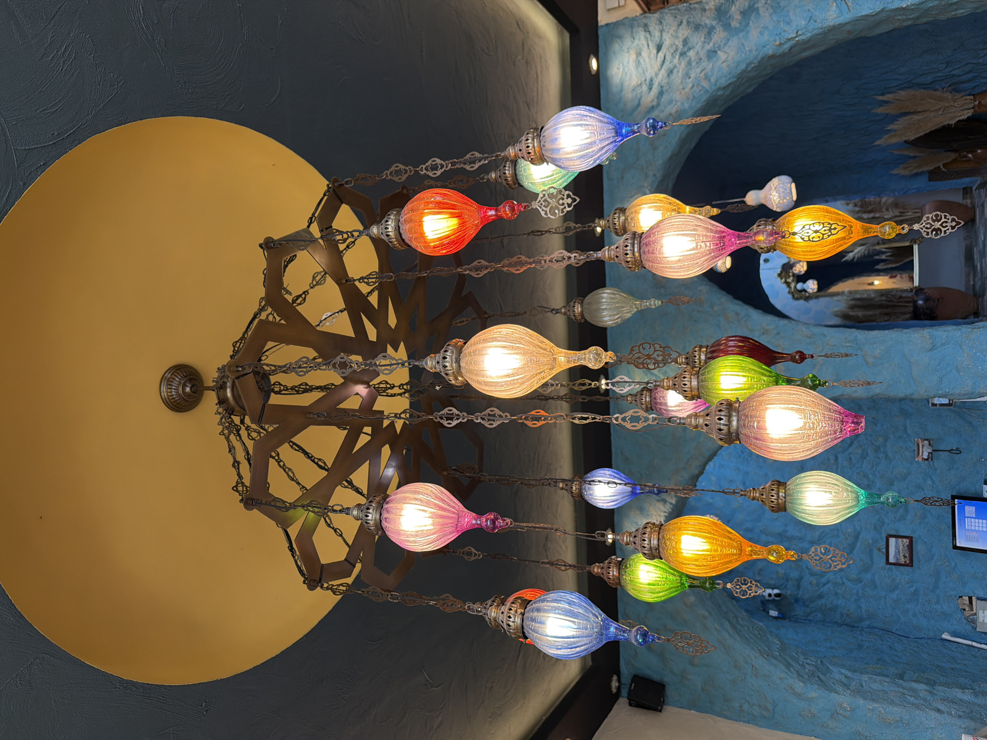 An ornate chandelier with numerous colorful, glass pendant lights hangs from a dark teal ceiling with a large, mustard yellow circular cutout. The lights, in shades of pink, blue, green, yellow, and orange, are illuminated and cast a warm glow. The chandelier itself is made of dark metal with intricate chain links and decorative elements. Below the chandelier, the environment appears to be a restaurant or cafe with textured, bright blue walls that form arched alcoves. In one alcove, a small framed picture hangs on the wall, and a computer monitor is visible on a counter below. Dried pampas grass in large ceramic pots adds a decorative touch to the scene. The overall aesthetic suggests a vibrant and culturally inspired setting.