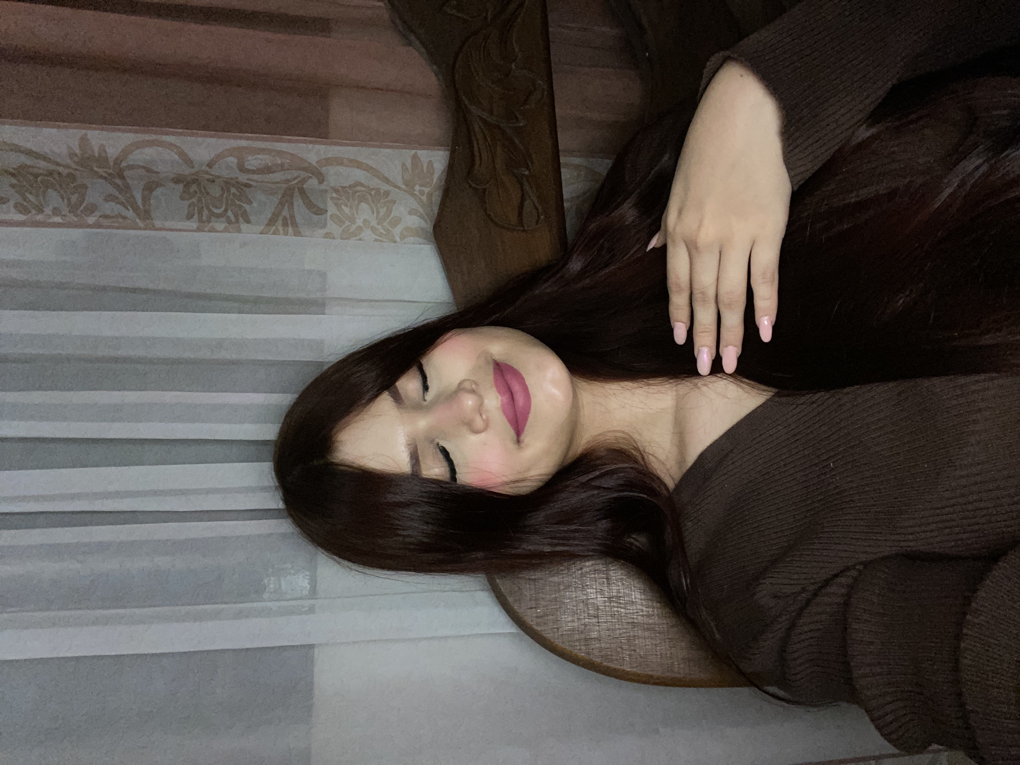 A woman with long, dark hair is lying down. She is wearing a brown sweater. Her face is visible, and she has pink lipstick on. Her hand is resting on her hair. She is on or beside a wooden chair or piece of furniture, which is adjacent to a wall or surface with a patterned edge or trim. The location is Juan Griego, Venezuela, although nothing in the image specifically indicates this.