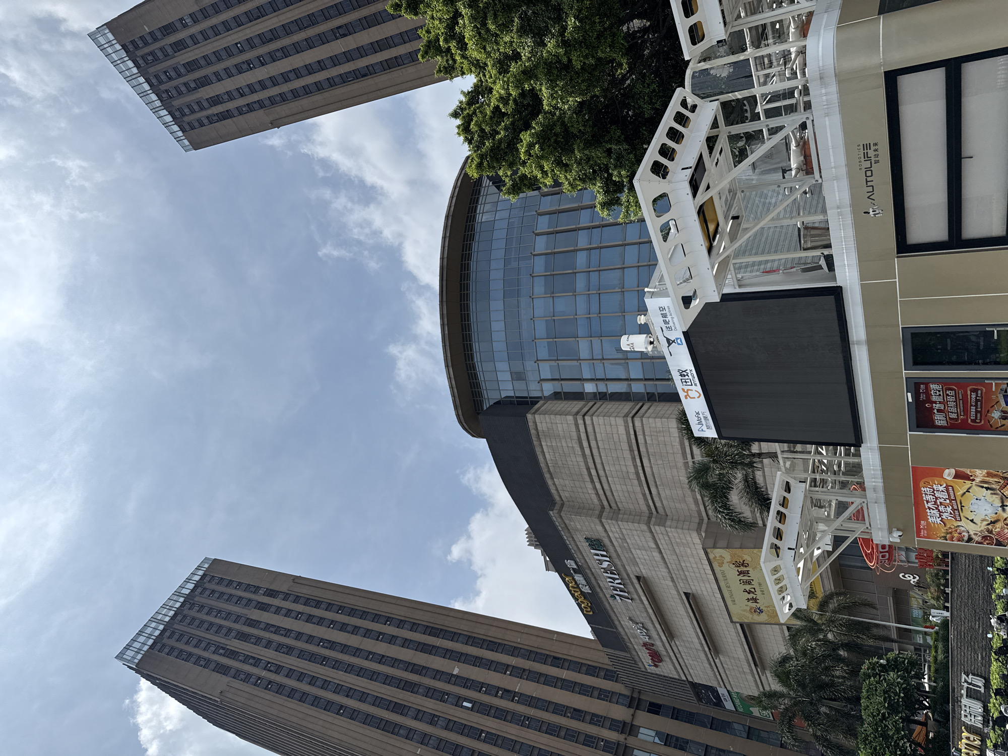 The image depicts a modern urban scene in Haizhu District, Guangzhou, China, featuring multiple tall buildings and a commercial complex under a partly cloudy sky.
In the left portion of the frame, a tall, slender skyscraper with a beige facade and numerous rectangular windows extends upwards, its top section featuring a glass parapet. A similar skyscraper, partially obscured by green trees, is visible on the far right, also topped with a glass parapet.
Centrally positioned below these towers is a multi-story commercial complex constructed with light-colored, possibly stone-clad, horizontal sections. Prominent signage on this complex includes "together 跃领荟" in orange and "7FRESH" in green and white. Below these, a large, golden-framed sign displays "PAILONGGE RESTAURANT 牌笼阁酒家" in red Chinese characters. A "CART 4D" sign is partially visible to the left. Further back, a large, circular building with a blue glass facade forms part of the complex. Palm trees are visible along the mid-ground, interspersed with the buildings.
In the immediate foreground, a white and beige mobile or fixed structure is present. Its left side features a large, dark digital display screen. The top of this structure consists of multiple white, framed bays or platforms. Text visible on the structure includes "ROBOTICS AUTOLIFE 智动未来" and above the display, the "美团" (Meituan) logo alongside "蚁蜂航空" (Antbee Aviation) with the English phrase "Delivering Airlines." Inside one of the open bays, a shovel or broom and a metal bucket are visible. Below this structure, red posters are displayed, one mentioning "美味不等待 外卖飞客来" and another referencing "保利广场 饭店" (Poly Plaza Restaurant). No identifiable people are clearly visible engaging in specific actions within the frame. The sky is predominantly blue with scattered white, wispy clouds.
