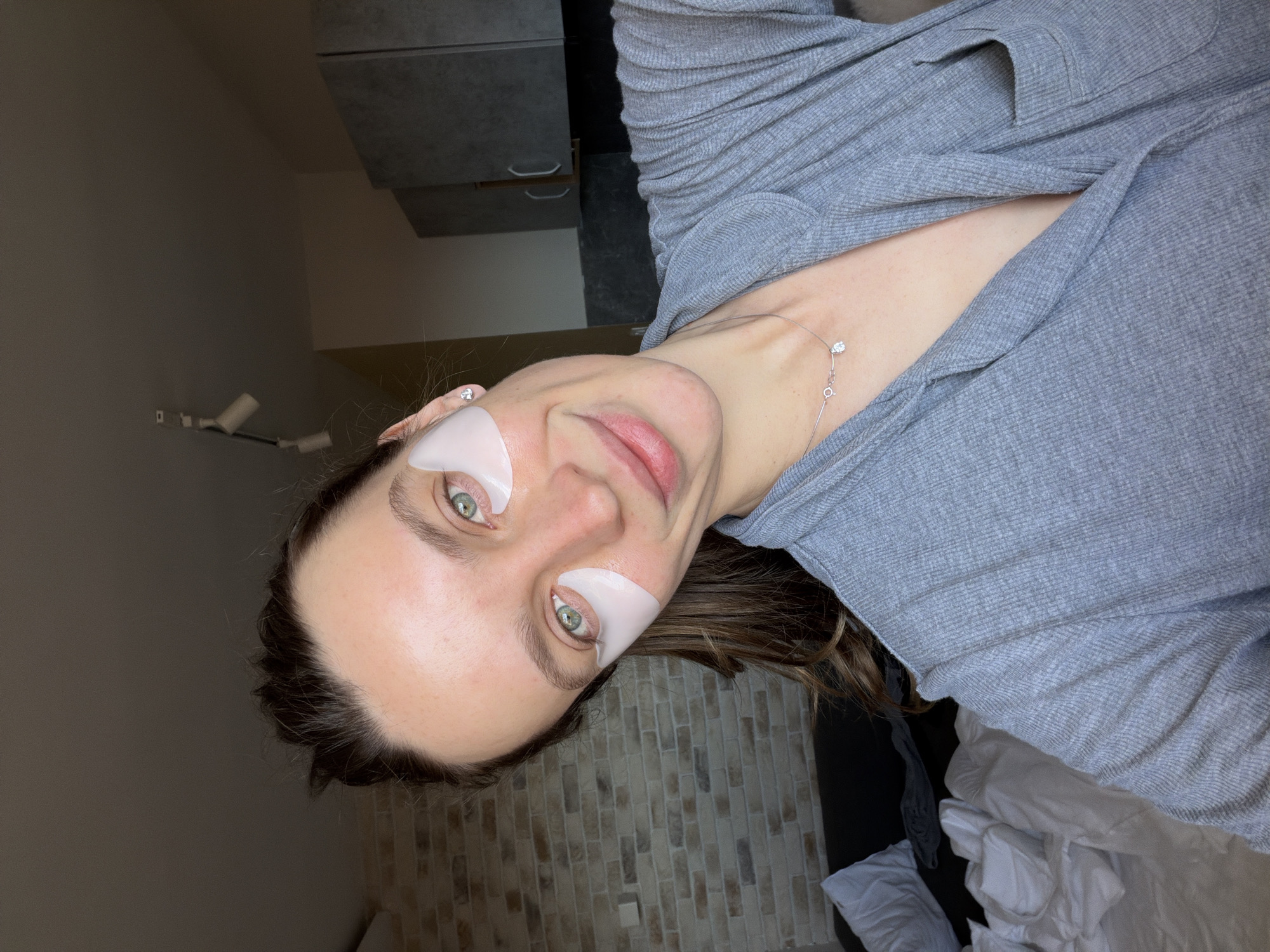 A woman wearing eye patches is lying on a bed. She is wearing a grey shirt and a necklace. Behind her is a wall with a brick pattern and a grey cabinet. The location is not discernible as Budva, Montenegro, based on the image provided.