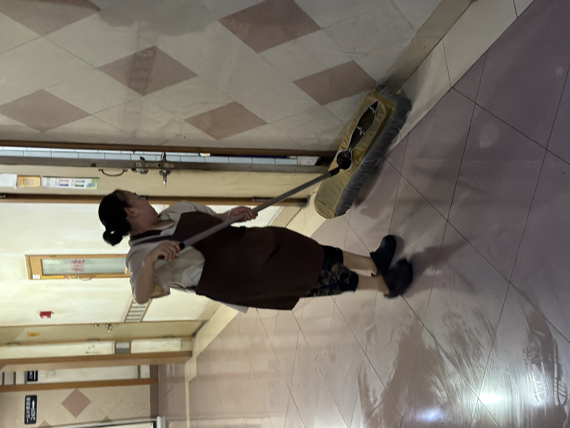 A person is mopping a tiled floor in a hallway. The person is wearing a dark apron over a light-colored shirt and patterned pants. They are using a wide mop with a yellow head. The hallway has light-colored walls with a diamond pattern and doors on either side. The floor tiles are a pale pinkish-brown color and appear to be wet and reflective. The scene appears to be in Yuzhong District, China, possibly in a hospital or other institutional setting, suggested by signage in the background.