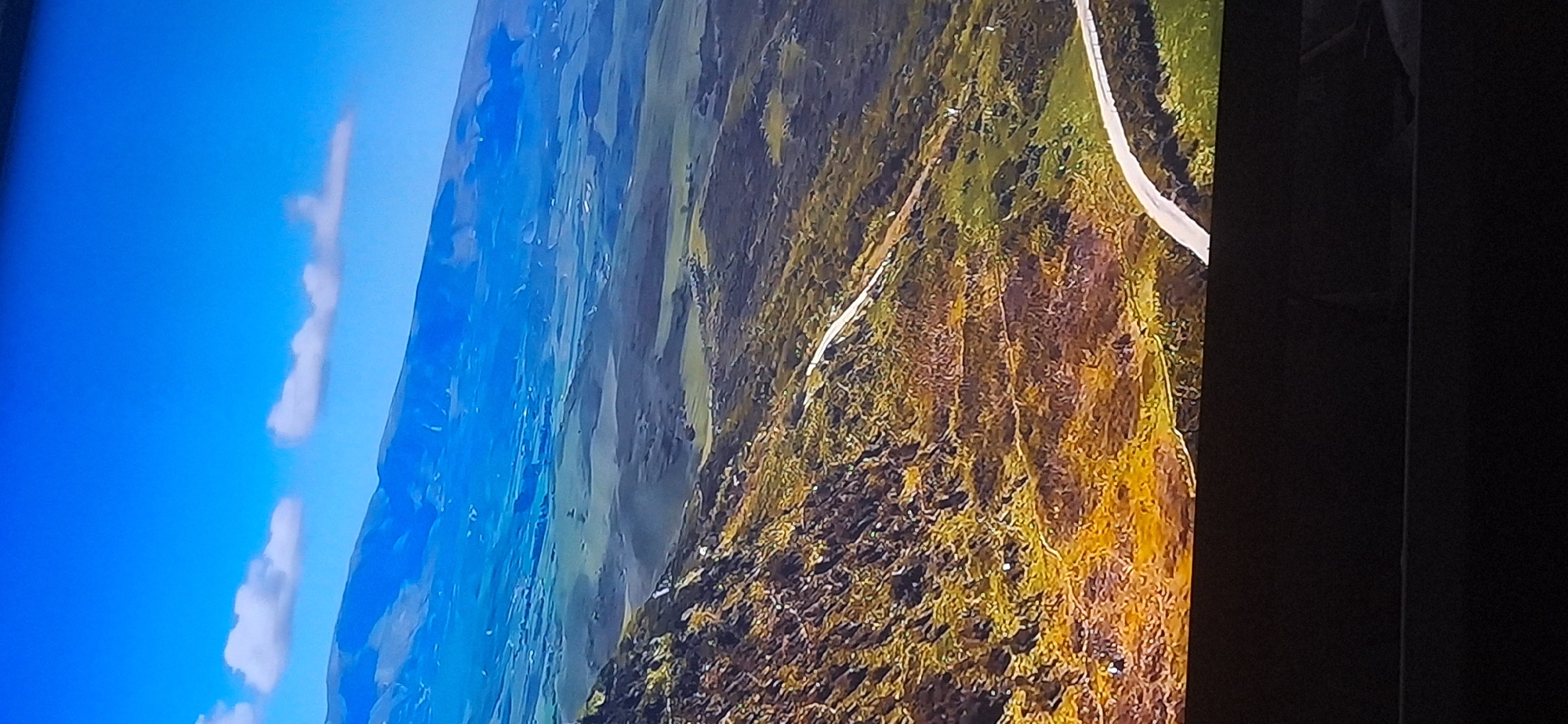The image shows a panoramic view of a mountainous landscape under a clear blue sky with a few scattered clouds. The foreground is dominated by a steep, rocky slope covered in dry, yellowish-brown vegetation and scattered patches of green. A winding road, appearing to be paved, carves its way up the slope and continues into the distance. Beyond the immediate slope, the terrain softens into rolling hills and valleys, painted in shades of green and brown. Further in the background, a vast, hazy mountain range stretches across the horizon under the bright blue sky. The scene appears to be a natural, undeveloped area, likely a rural or mountainous region. There are no discernible buildings, structures, or any signs of urban development in the image. There are no people or animals visible. The image does not depict the city of Bila Tserkva, Ukraine.