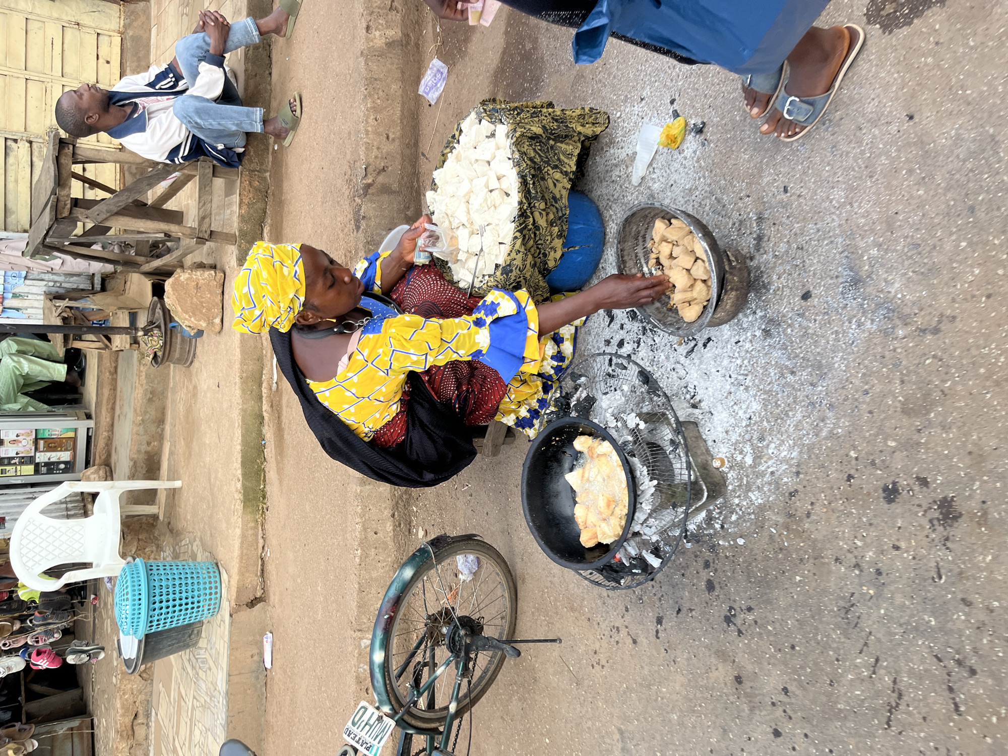 In the city of Bukuru, Nigeria, a woman is seen frying food in a black pan placed over burning coals. She is wearing a bright yellow and blue patterned dress and a yellow headscarf. In front of her, to the right, is a large bowl filled with white cubes, possibly cheese or tofu, draped with a patterned cloth. Next to this bowl, a man in a white jacket and blue jeans is seated on a wooden bench, looking to the side. To the woman's left, a bicycle with a license plate reading "PLATEAU MUHO" is parked. The ground is paved with concrete and there are some scattered white powders and debris. In the background, there are stalls with various items, including shoes and products on shelves, and a white plastic chair. The overall scene depicts a busy street-side market or food vendor stall.