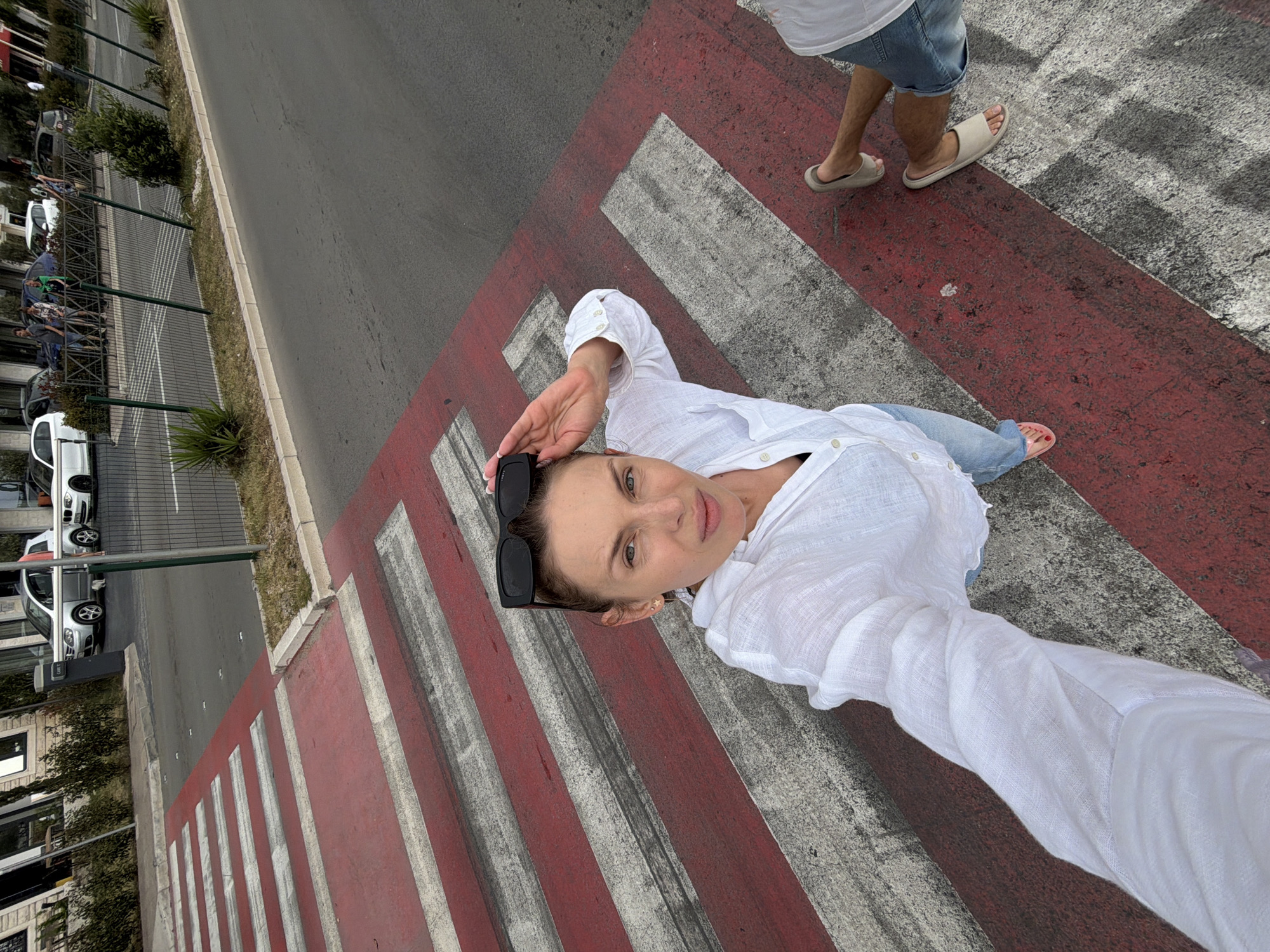 A woman takes a selfie on a vibrant red and white striped pedestrian crossing in Budva, Montenegro. She is positioned diagonally across the frame, looking directly into the camera with a calm expression. She wears a white, loose-fitting long-sleeved shirt and light blue jeans. Dark sunglasses are perched on top of her head. Her right hand is raised, partially shading her forehead. One of her feet, with pink painted toenails, is visible near the bottom of the frame.
To the right of the woman, the lower legs and feet of another person are visible, wearing denim shorts and light-colored slip-on sandals, suggesting they are also on the crossing.
The crossing itself is painted on an asphalt road. To the left, a multi-lane street runs parallel, bordered by a concrete curb, a narrow strip of grass, and a metal fence. Several cars are parked along the fence line. Beyond the street, buildings with multiple stories and more greenery are visible. The scene is brightly lit by daylight, indicating clear weather. No visible text is present in the image.