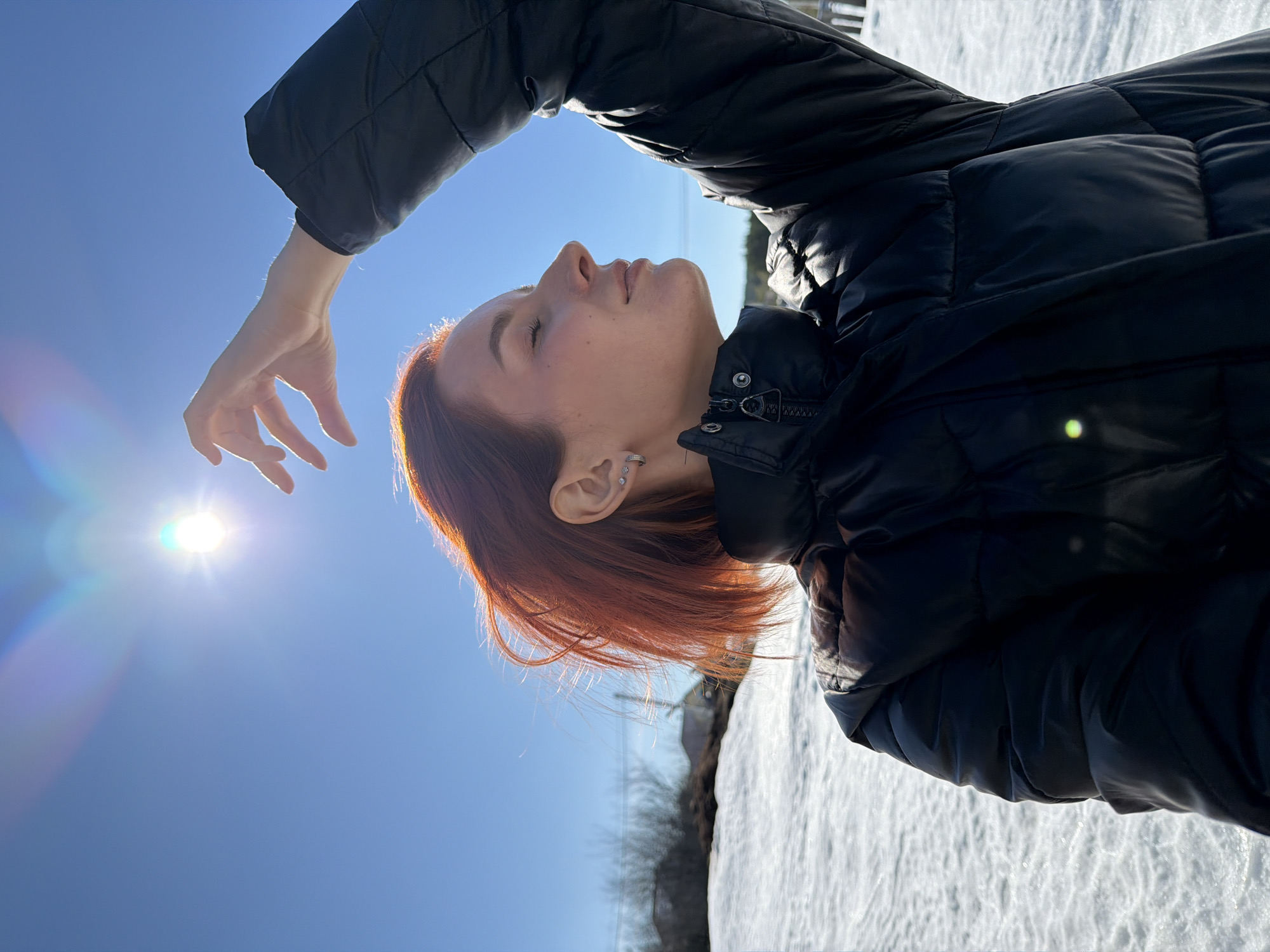 A female individual is depicted outdoors in Chaikovsky, Russia, under a clear blue sky with a bright sun positioned in the upper left quadrant, exhibiting lens flare. The subject, appearing from the chest up, has short, reddish-orange hair, slightly windswept to her left. She wears a black puffer jacket. Her right arm is raised with fingers splayed, positioned to partially shield her face from the sun. Her head is tilted slightly upward and to the left, with her eyes closed and a subtle smile on her lips. Multiple earrings, including a hoop and studs, are visible in her left ear. The background features a snow-covered landscape in the lower half of the frame, with distant, bare trees and indistinct structural elements visible near the horizon. The scene indicates a sunny winter or early spring day.