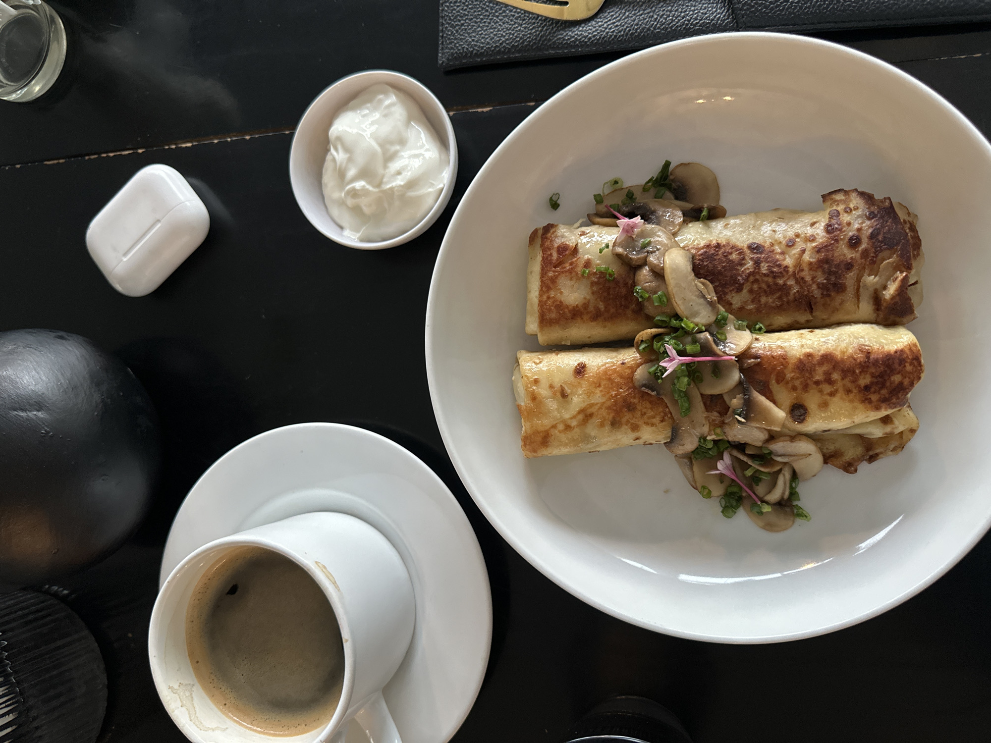 An overhead, eye-level shot captures a meal at a dark table, possibly in Kuta Selatan, Indonesia, featuring a plate of syrniki garnished with mushrooms and chives, a white cup of coffee with a saucer, a small bowl of sour cream, and a white wireless earbud case. The syrniki are golden brown and cylindrical, topped with sliced mushrooms and finely chopped green chives, with delicate pink edible flowers. The coffee is dark and steaming, in a simple white mug. The sour cream is thick and white, in a small ceramic dish. The earbud case, a common modern accessory, sits to the right of the coffee. The dark table surface suggests a restaurant or cafe setting, with the overall composition suggesting a breakfast or brunch.