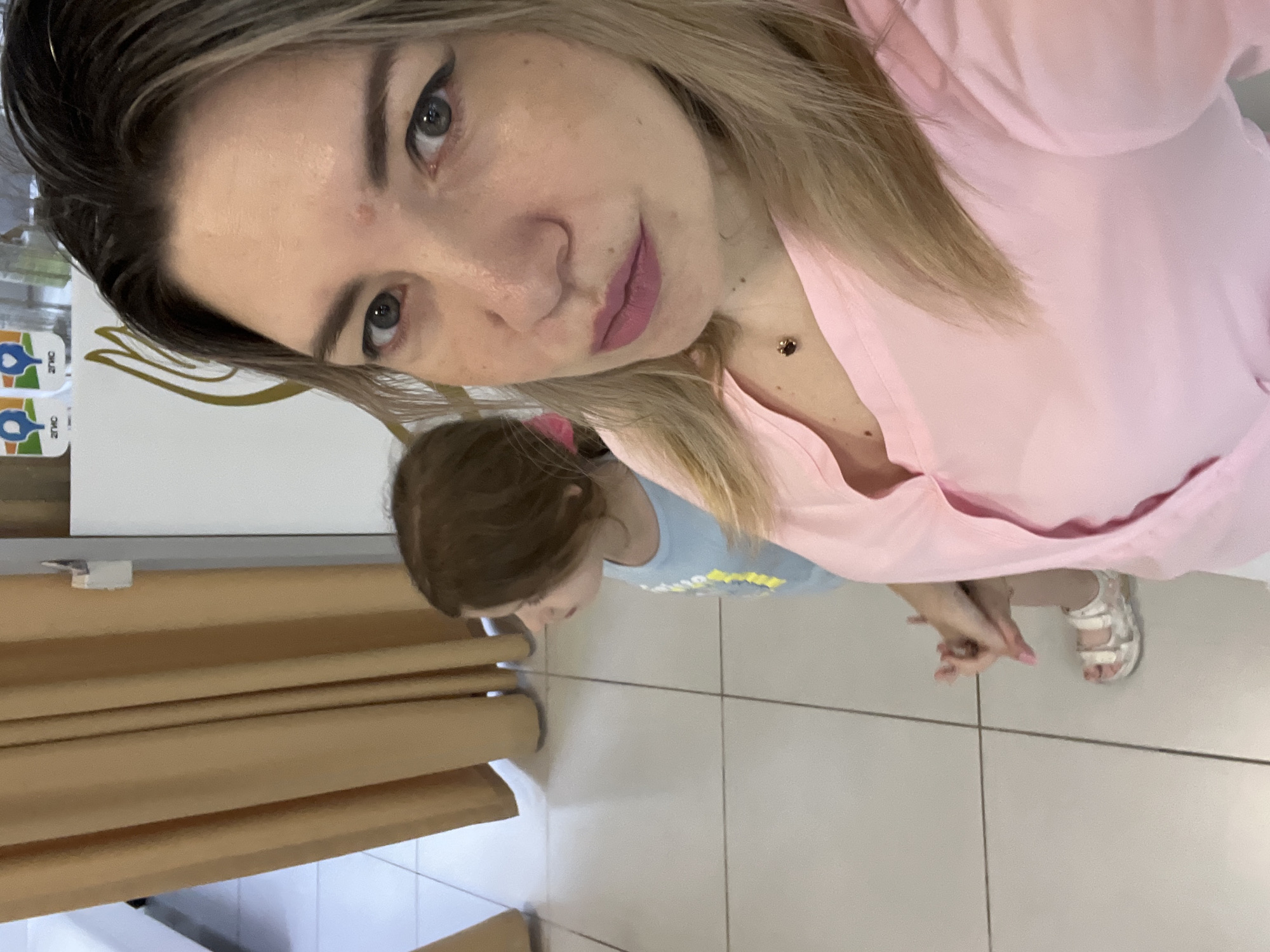 In Sochi, Russia, a woman with blonde hair and makeup is taking a selfie. She is wearing a pink shirt and a small dark pendant necklace. To her left, a young girl with brown hair is looking away from the camera. She is wearing a light blue t-shirt and white sandals. The background shows a wall with a light-colored curtain and a white surface with some logos and bottles. The floor is tiled with a light color and dark grout lines.