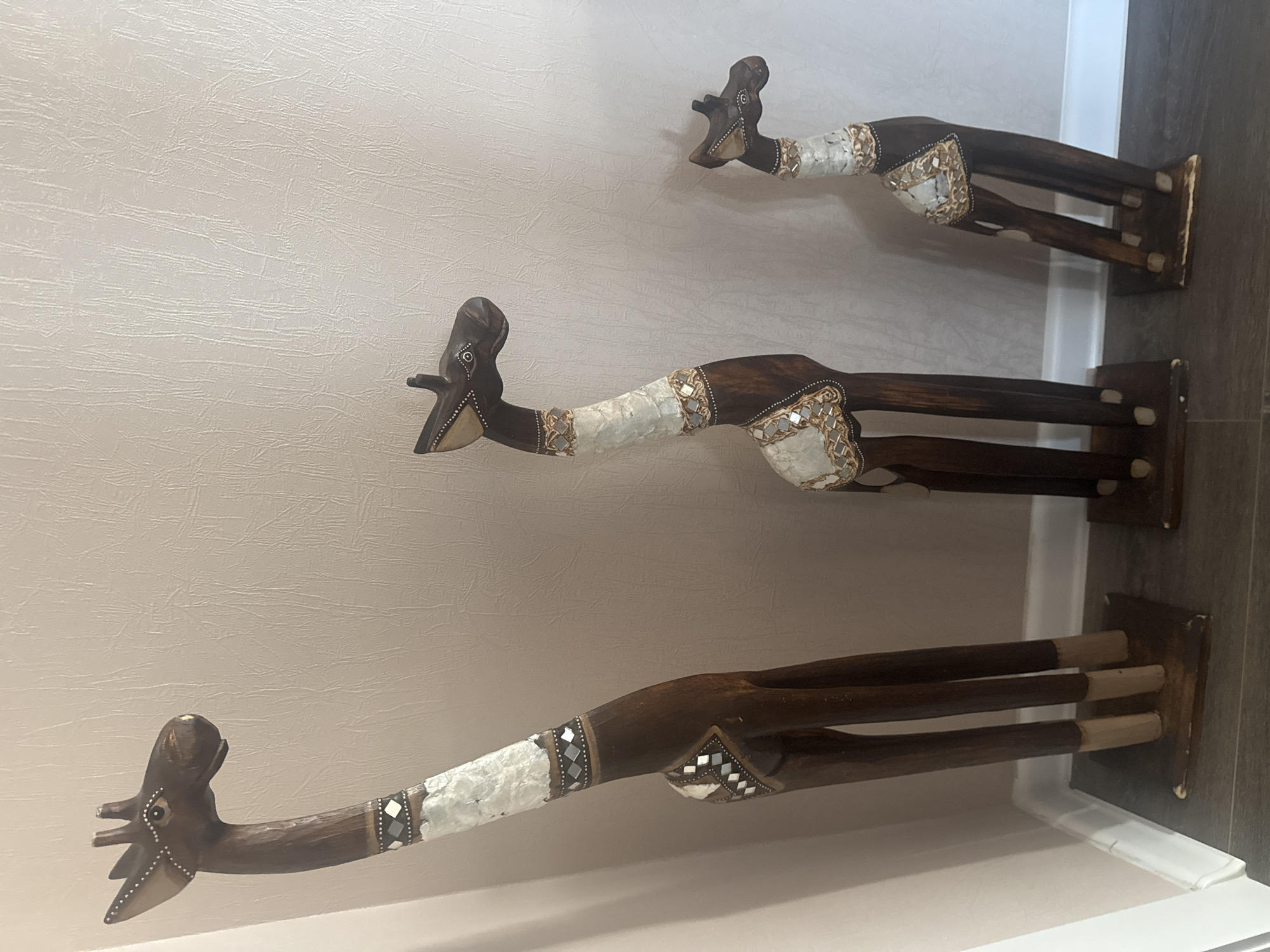Three decorative wooden giraffe figurines of varying heights stand on a dark wooden floor against a textured light beige wall. The giraffes are dark brown and adorned with inlaid patterns of white material, small mirrored squares, and decorative beads around their necks and upper bodies. The largest giraffe is on the left, followed by a medium-sized one in the center, and the smallest on the right. The bases of the giraffes appear to be dark wood squares. The environment suggests an indoor setting, likely a home or retail space. There are no people or actions depicted in the image. The location is not specified within the image itself.