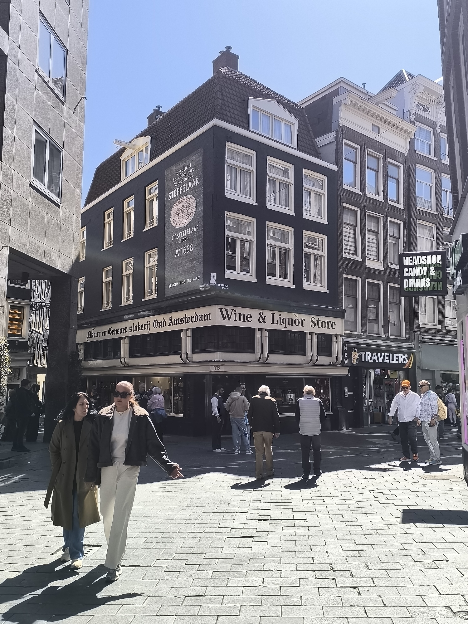Here is a description of the image:
The image captures a street scene in Amsterdam, Netherlands, showcasing architectural elements and pedestrian activity under a bright, sunny sky. The focal point is a corner building advertising "Wine & Liquor Store" with the name "Likeur en Genever stokerij Oud Amsterdam" and "C.T. STEFFELAAR Leiden A°1658". The building has a dark facade with white-framed windows.
Several pedestrians are present. In the foreground, two women are walking along a paved street with a grid pattern. One woman wears a long green coat over blue jeans, while the other is wearing white pants and a dark jacket. To their right, further down the street, are other people strolling. A sign reading "HEADSHOP, CANDY & DRINKS" is visible next to another shop sign that reads "TRAVELERS".
The environment includes buildings with varying architectural styles and heights. Some are dark-colored, while others are lighter in tone. The overall impression is that of a busy urban street with a mix of commercial and residential structures.