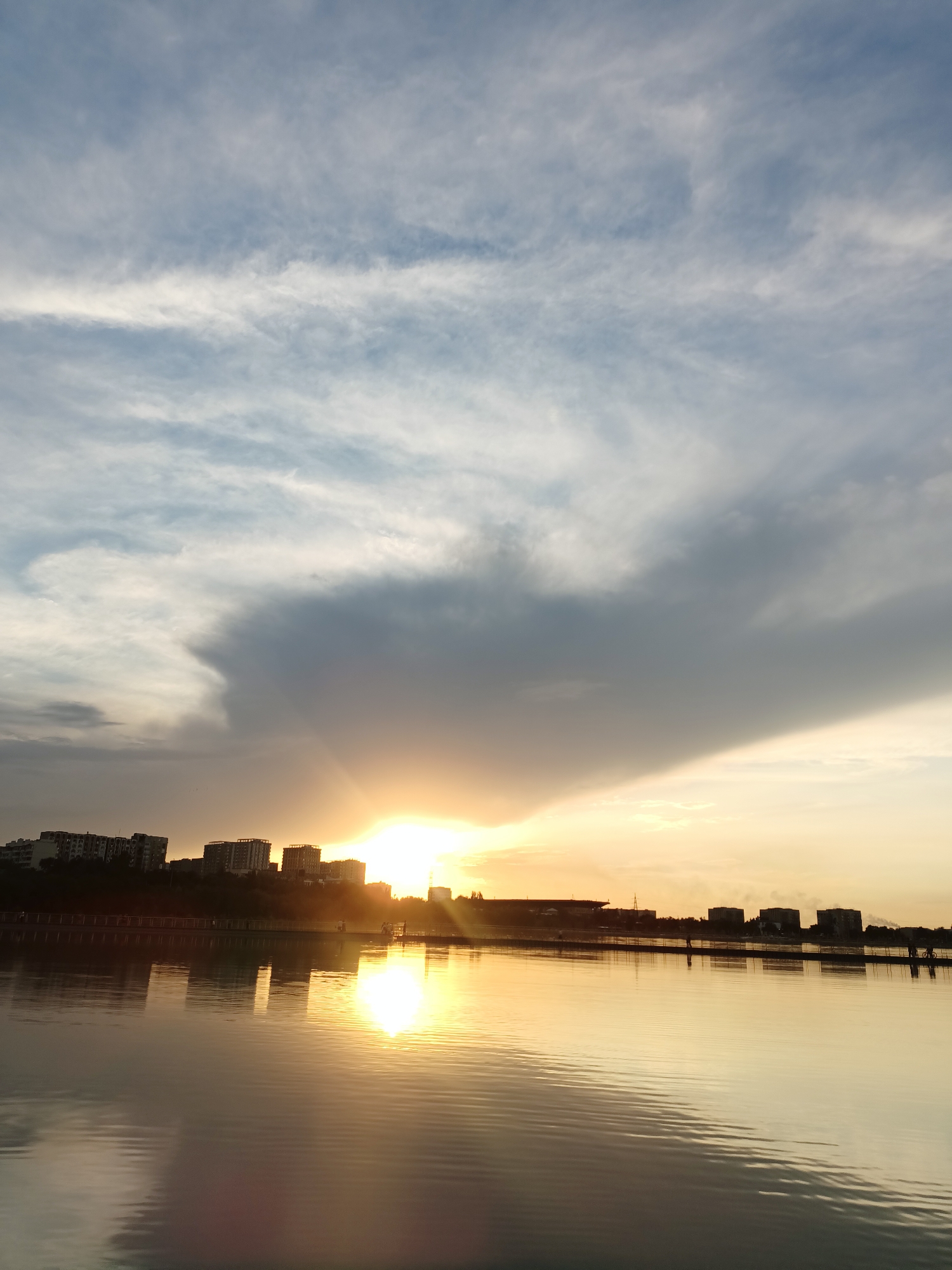 The image depicts a tranquil scene at sunset over a body of water in Almaty, Kazakhstan. The sky is filled with a dramatic display of clouds, with wispy white formations near the top and larger, darker clouds gathering towards the right, obscuring parts of the setting sun. Rays of sunlight pierce through the clouds, casting a warm, golden glow across the sky and reflecting on the water's surface.
In the background, a city skyline is silhouetted against the vibrant sky. Numerous buildings of varying heights are visible, suggesting an urban environment. A pier or walkway extends across the water in front of the buildings, with a few indistinct figures of people visible along its length. One person appears to be on a bicycle. The water itself is calm, with gentle ripples creating distorted reflections of the buildings and the sky. The overall impression is one of serene natural beauty enhanced by the urban landscape.