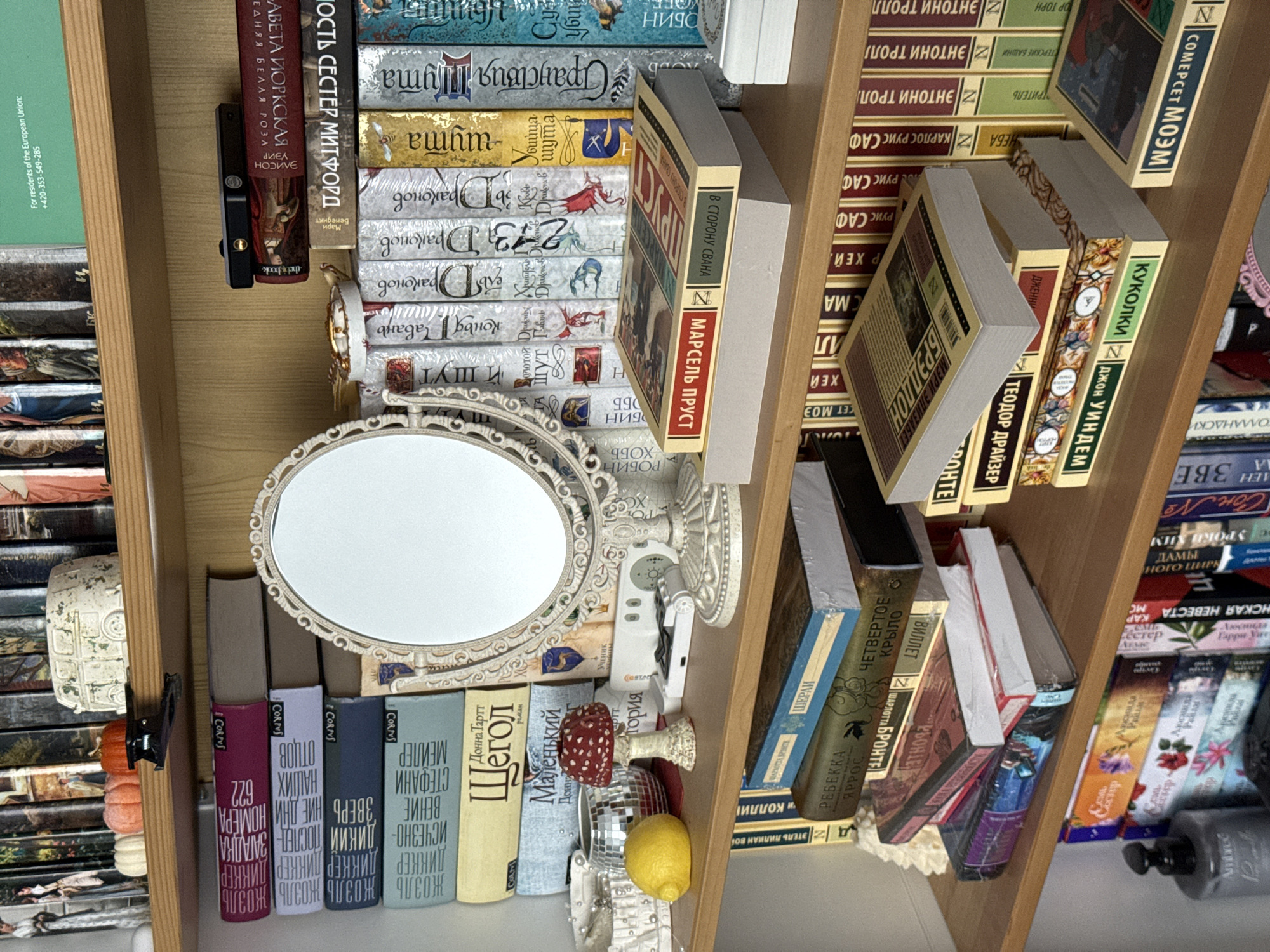This image captures a section of three light-brown wooden bookshelves against a light-colored wall, characteristic of an indoor residential or study environment in Krasnodar, Russia. The shelves are densely populated with books and various decorative objects.
On the uppermost shelf, from left to right, are a small white ceramic pumpkin, an orange ceramic pumpkin, a black hair claw clip, and a small white ceramic sculpture of a Volkswagen T1 van. Multiple books with predominantly Russian titles are arranged behind these items.
The middle shelf prominently features an ornate, antique-style oval standing mirror with a cream-colored frame. To the left of the mirror, a small white basket holds white beaded accessories, a yellow lemon, a miniature disco ball, and a red-spotted ceramic mushroom figurine. A collection of horizontally stacked books, including Russian editions by Joel Dicker and Donna Tartt, is visible. To the right of the mirror, a small white rectangular electronic device and a black rectangular electronic device rest on stacks of books. A Russian edition of Marcel Proust is placed face down on a stack of grey books. Other books with varied spine designs and Russian text line this shelf, one bearing a green stripe with text "For residents of the European Union: +420-353-549-285."
The lowermost visible shelf contains additional books, arranged both vertically and in stacks. On the far left, a dark-colored bottle with a pump dispenser, labeled "Ambre," stands next to a white ceramic Christmas tree-shaped decoration. Visible book titles include works by Anthony Trollope, Somerset Maugham, John Updike, and Theodore Dreiser.
No individuals or active interactions are present; all objects are stationary.