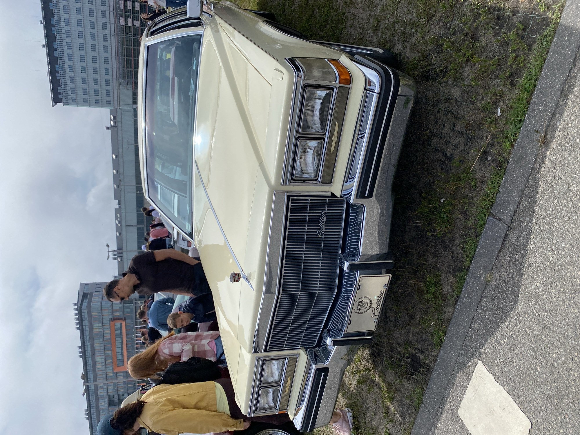 In Gdańsk, Poland, a pale yellow Cadillac is parked on a grassy area next to an asphalt path, with a large, modern building with glass windows and a prominent orange section in the background. A group of people are gathered around the car, some looking at it. The front of the Cadillac, featuring a prominent chrome grille and rectangular headlights, is the main focus. A license plate with the Cadillac crest and lettering is visible. The sky is overcast.