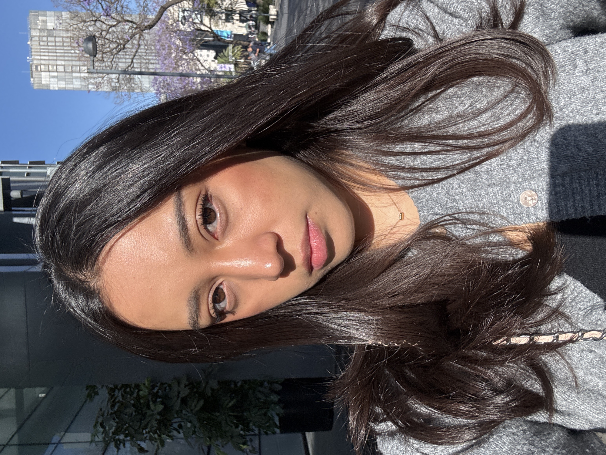 A young woman with long, dark hair and brown eyes is photographed outdoors. Her face is angled slightly towards the camera, with a light source illuminating her features. She is wearing a grey knit sweater and a delicate gold necklace. In the background, trees with purple blossoms are visible, along with buildings characteristic of an urban setting. Based on the distinctive jacaranda trees and architecture, the location is likely Mexico City, Mexico.