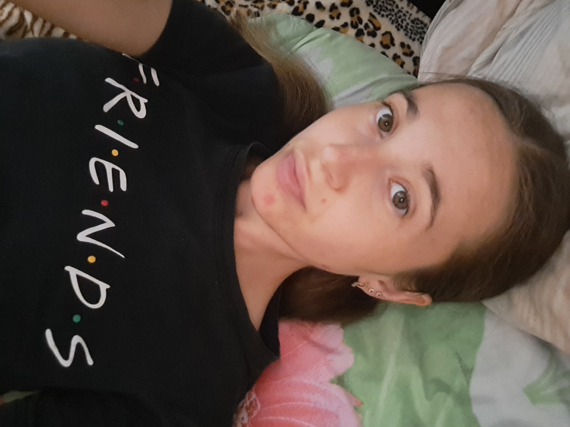 A young woman with light brown hair lies on her side in bed, looking at the camera. She is wearing a black t-shirt with the word "FRIENDS" printed in white letters, each dotting one of the 'i's with a different color. Her head rests on a pillow with a light green and white patterned cover. A leopard-print blanket is visible to her left. The background is a close-up of a bed with a floral patterned duvet, in shades of pink and green. The image is taken from a slightly high angle.