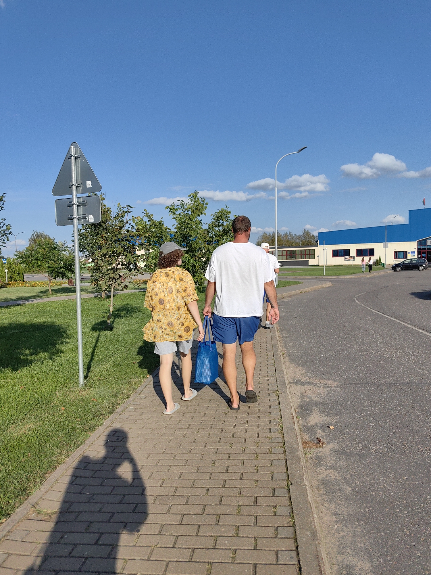 Two people walk along a paved sidewalk beside a grassy area and a road. The person in front is wearing a yellow shirt with a sunflower print and gray shorts. They are carrying a blue tote bag. The person behind them is wearing a white t-shirt and blue shorts. A shadow of a third person, holding a camera, is cast on the sidewalk. In the background, there is a modern building with blue and white accents, a lamppost, trees, and a traffic sign. The sky is clear blue with some white clouds. The location is the perimeter of a facility in the city of Myadzyel, Belarus.