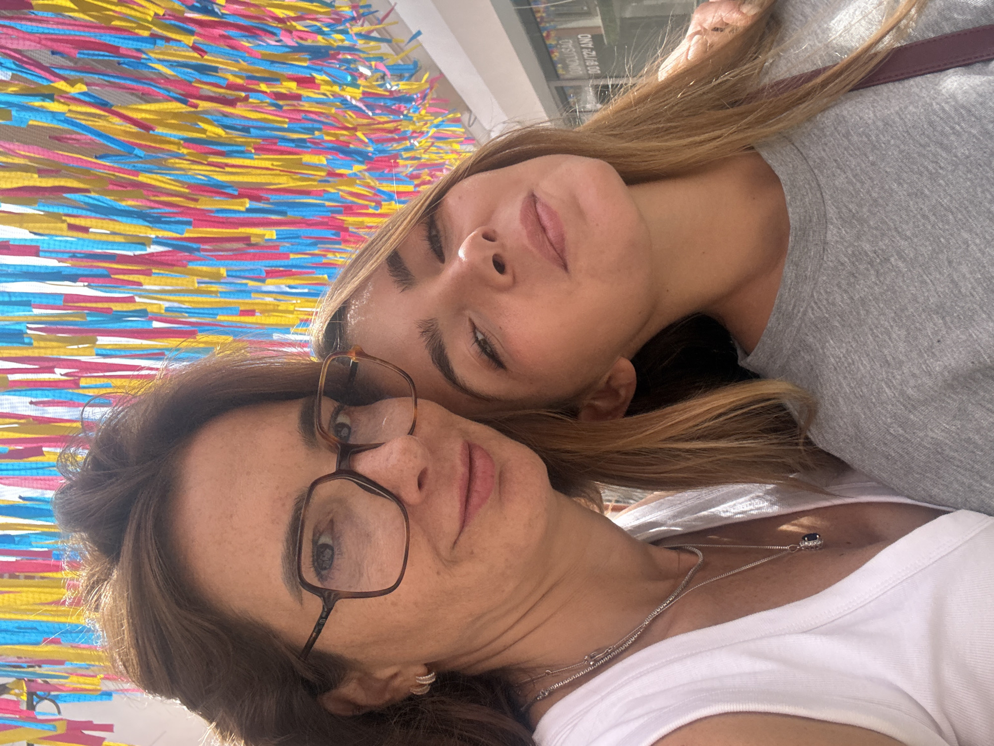Two people, an adult female and a young female, are taking a selfie outdoors. The adult female is in the foreground on the left, wearing glasses, a white tank top, and necklaces. The young female is to her right, wearing a grey t-shirt. They are both looking towards the camera. Behind them, a colorful display of hanging streamers in yellow, pink, and blue is visible against a bright sky. In the background, a storefront with signage is partially visible. The scene appears to be in the city of Torres Vedras, Portugal.