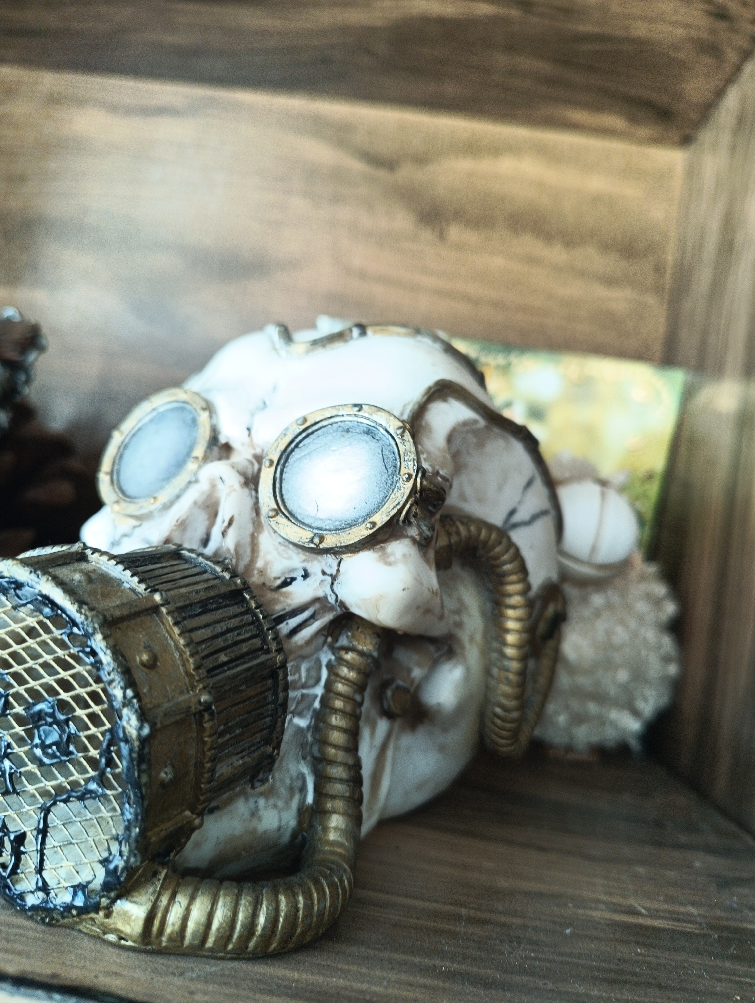 A close-up, eye-level shot shows a steampunk-themed skull sculpture with goggles and a respirator mask. The skull is predominantly white with gold accents. The goggles have round, reflective lenses and a metallic gold rim. A gold-colored, ribbed tube extends from the side of the skull, connecting to the respirator mask, which features a circular grille with a diamond pattern and gold trim. The skull is positioned on a wooden surface, and a wooden shelf or frame is visible behind it, creating a shadowbox effect. The background includes a hint of a colorful, possibly floral, pattern on a piece of paper or fabric. The lighting is soft, with some bright highlights on the lenses. The scene appears to be indoors, possibly on a shelf or display in Karnobat, Bulgaria, given the context of the query.