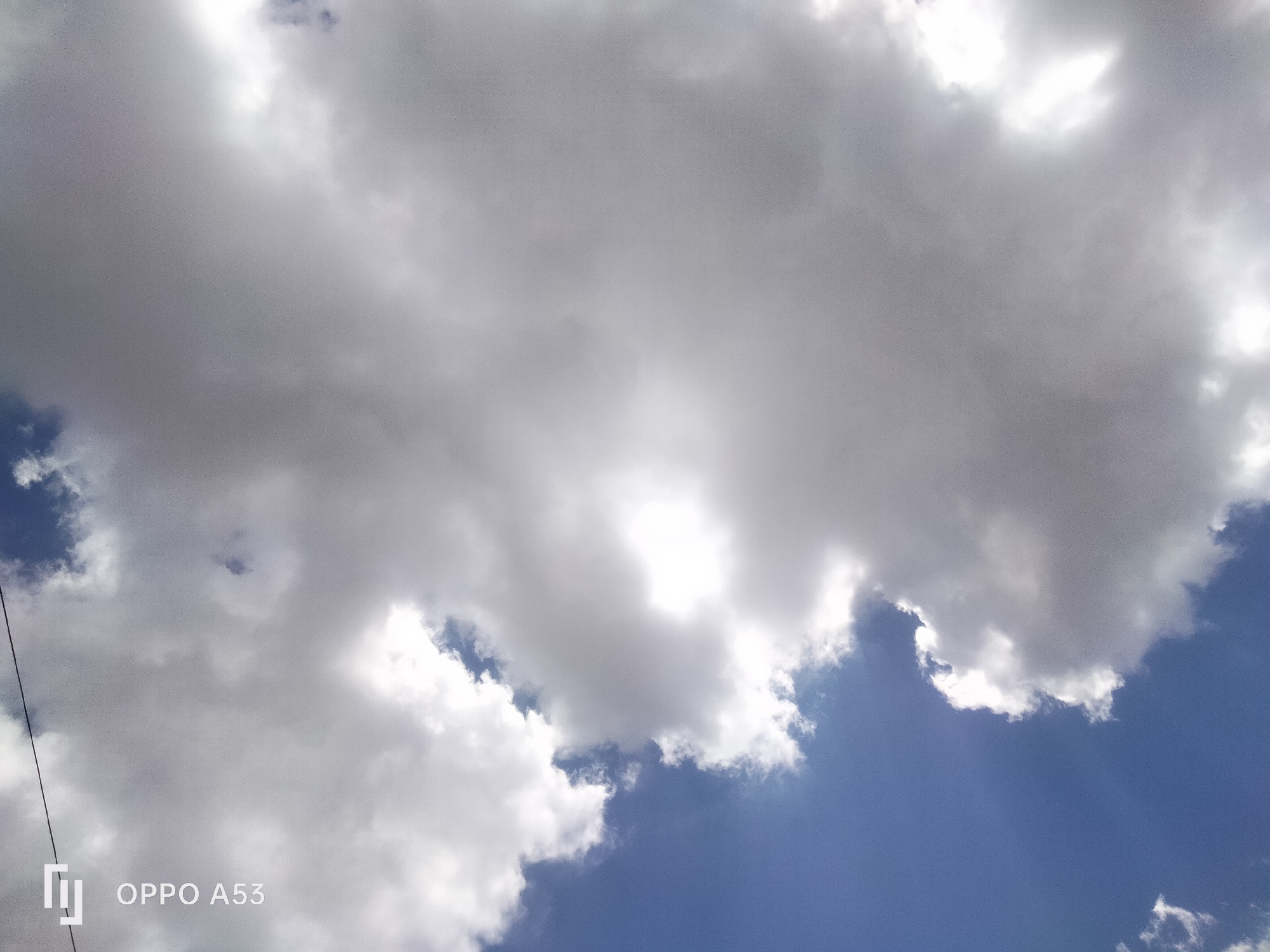 The image depicts a sky filled with cumulus clouds, some appearing bright white and illuminated by sunlight, while others are darker gray, suggesting varying thickness or moisture content. Patches of clear blue sky are visible between the clouds. In the lower left corner, a thin, dark, vertical line, likely a power line or cable, is present. Overlayed on the image in the lower left is a white watermark displaying a stylized "U" shape next to the text "OPPO A53," indicating the device used to capture the photograph. The scene is a natural outdoor environment, likely a daytime sky. The location in Badung, Indonesia, suggests this could be a common sky view over residential areas, agricultural land, or coastal regions within the regency. There are no people or explicit objects other than the clouds and the watermark.