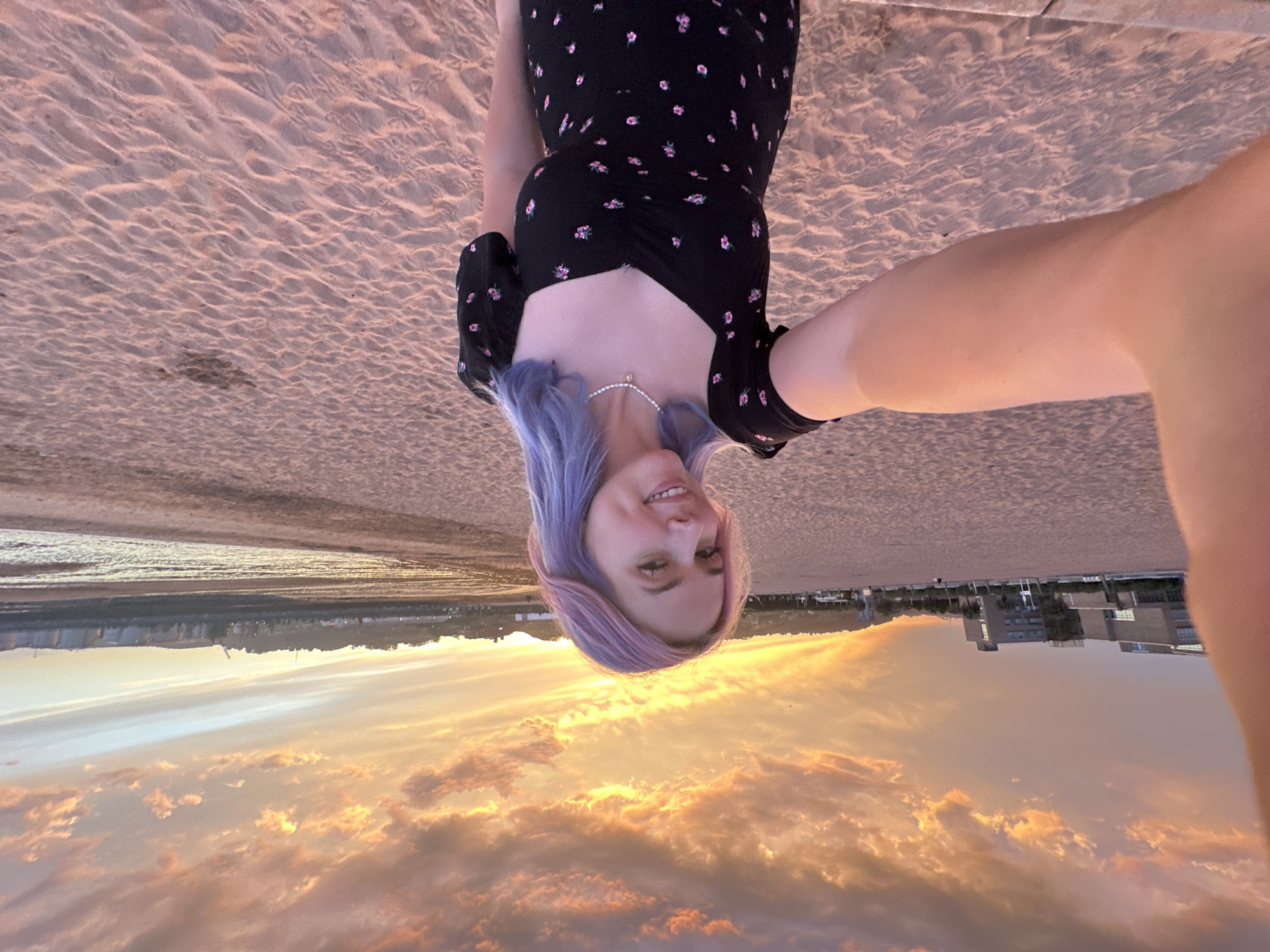 A young woman with purple and pink hair smiles at the camera while holding her phone to take a selfie. She is wearing a black dress with a floral print and a pearl necklace. The woman is standing on a sandy beach, with the ocean and a cityscape in the background. The sky is filled with dramatic clouds, colored orange and pink by the setting sun. The scene appears to be the El Muelle beach in Gijón, Spain, which is located near the city's harbor and marina, with buildings and a hill visible in the distance.