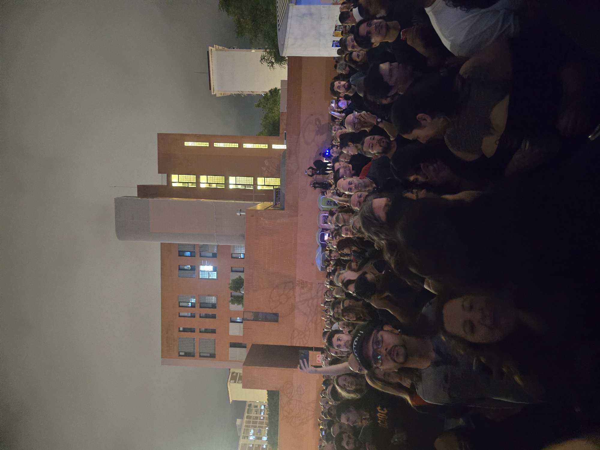 This nocturnal image captures a densely packed outdoor gathering of predominantly adult individuals in Cornellà De Llobregat, Spain. In the foreground and midground, a large crowd of people, mostly dressed in dark, casual attire, faces forward, suggesting an unseen focal point beyond the frame. Several individuals are visible holding smartphones above their heads, indicating recording or photography. Notable individuals include a person in the lower left wearing an AC/DC shirt and another in the lower center with glasses and a goatee.
In the mid-background, a large, multi-story building constructed of reddish-brown brick dominates the scene, featuring numerous rectangular windows, some of which are illuminated with warm yellow light. A tall, slender, cylindrical structure with a grey, textured surface rises centrally between segments of this building. To the right of the brick building, a white, multi-story building is visible in the distance. A lower brick wall, adorned with faint graffiti, separates the crowd from the main building complex.
On the left side of the mid-background, two portable toilets, one blue and one green, are visible. To the right, a white temporary structure stands. Elevated figures can be observed on a platform or wall in the background, near the red brick building. The sky is overcast and grey, indicating artificial light reflecting off clouds in a nighttime setting. Blue light is discernible near the portable toilets, possibly from event lighting.