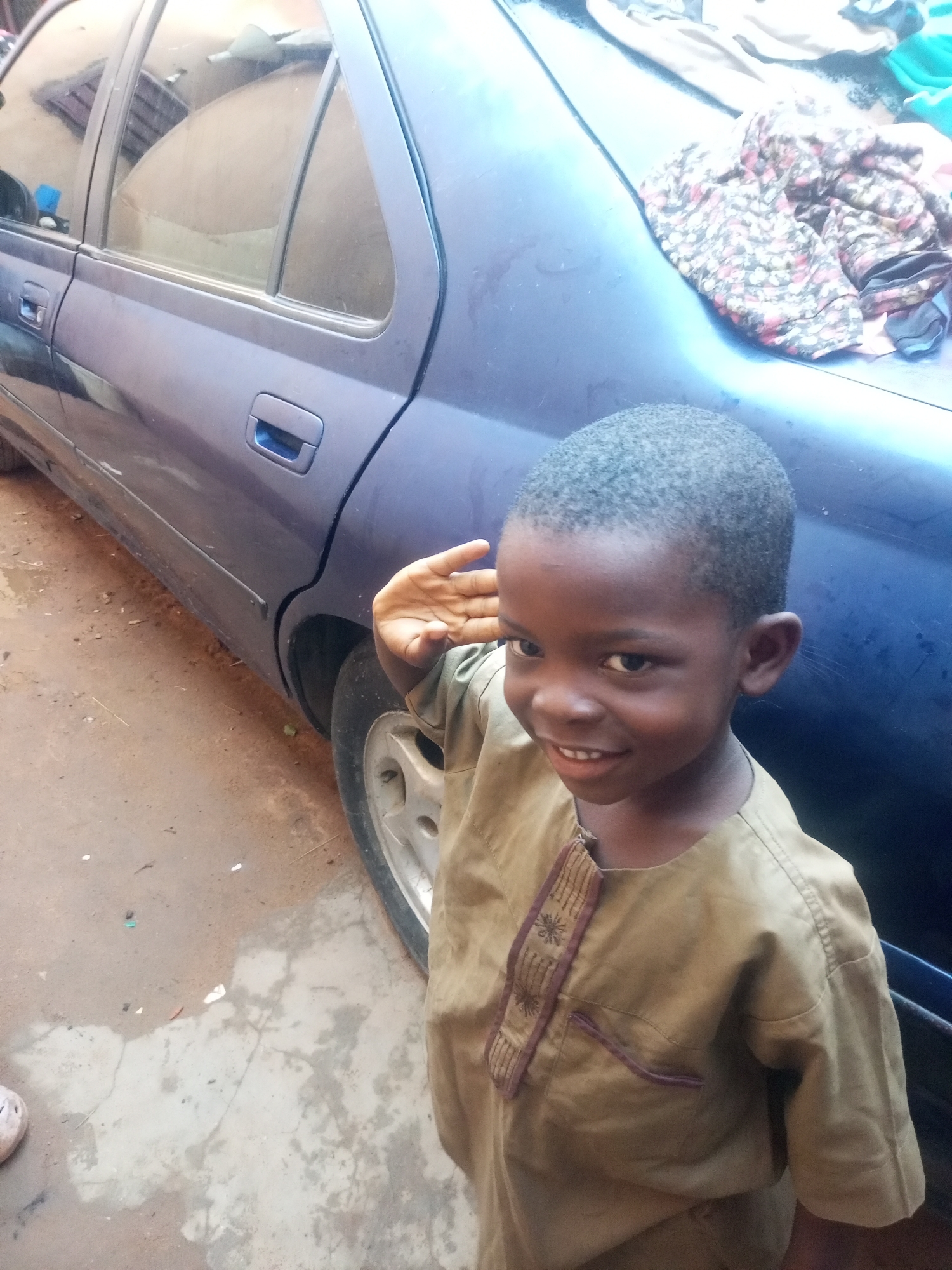 A young boy with dark skin and short, dark hair is standing next to the rear passenger side of a dark blue car. The boy is wearing a light brown, short-sleeved traditional garment with purple embroidered trim on the placket and a chest pocket. His left hand is raised to his forehead in what appears to be a salute. He is smiling at the camera. The car is parked on a dirt and concrete surface, likely a street or yard, in a dusty environment. The background shows the side of the car, including a rear door handle and part of a wheel. Some folded clothing, appearing to be a floral print fabric and a light blue garment, is piled on the trunk of the car. The scene is lit by natural daylight. The context of Tempure, Nigeria is provided based on external information.