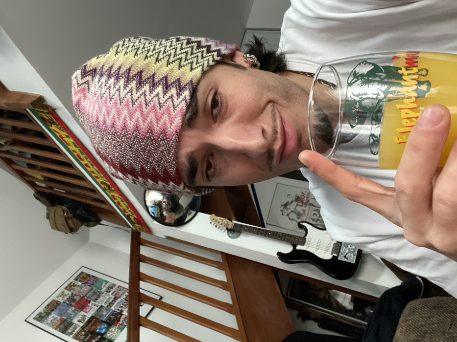 An indoor scene in Plaisir, France, depicts a young adult male from the chest up, positioned slightly to the right of the frame, looking directly at the camera with a slight smirk. He wears a multi-colored knitted beanie with a zigzag pattern in shades of pink, purple, yellow, and beige, and a white t-shirt. His dark hair is visible beneath the beanie, and he has a thin mustache and light stubble. A silver-toned chain-link earring is visible on his left ear. He holds a clear glass filled with an orange liquid, likely juice, in his right hand, with his index finger extended upwards beside the glass. The glass features a green elephant logo and yellow text reading "Elephantman" or similar.
The background reveals a residential interior. To the left, a wooden staircase with brown railings ascends upwards to the right. A framed collage or poster containing multiple smaller images is visible on the white wall near the stairs. Above the upper section of the stairs, a brown backpack hangs, and a fabric strip with red, yellow, and green horizontal stripes is draped along the banister. A circular convex mirror is mounted on the white wall below the banister. Below the mirror, a black electric guitar with a white pickguard is vertically mounted on the wall. A framed image is partially visible behind the guitar. A dark, possibly upholstered furniture piece is present in the lower-left foreground.