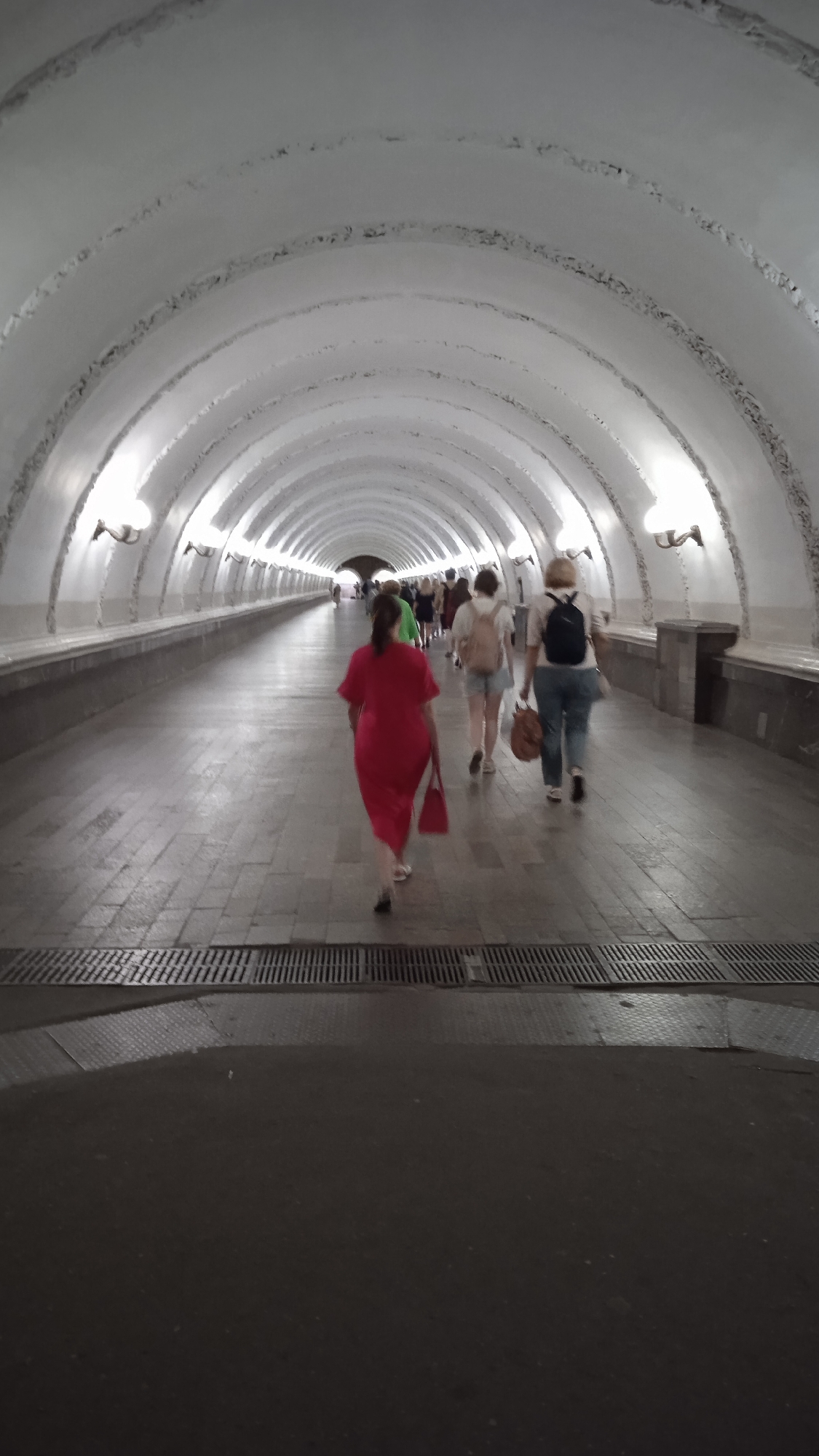 A long, white, arched tunnel with lights on the walls and people walking away from the camera. The floor is paved with grey tiles and has metal grates running across it. In the foreground, a person in a red dress is walking away, carrying a red bag. Further down the tunnel, other people are walking in the same direction, some carrying bags. The tunnel appears to be an underground passage, possibly a metro station or pedestrian underpass.