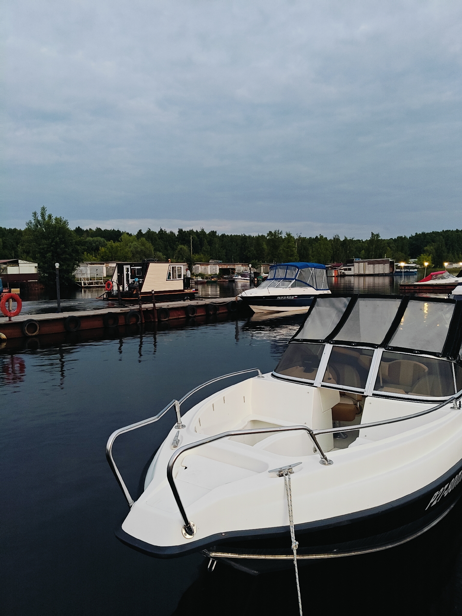 The image depicts a marina scene at dusk, with several boats docked along a wooden pier. In the foreground, a white motorboat with a black hull is partially visible. The boat has a windshield with dark framing and chrome railings. The water is dark and reflects the dim light.
Further along the pier, there are other boats, including a blue and white motorboat with a blue canopy. A houseboat, painted beige with white trim and a dark roof, is also present, with a person standing on its deck. In the background, a treeline of dark green coniferous trees stretches across the horizon, behind a row of small buildings and structures. The sky is overcast with a pale, diffused light.
The location is identified as a marina in Chaikovsky, Russia. The overall atmosphere is serene and quiet, typical of a harbor at the end of the day.