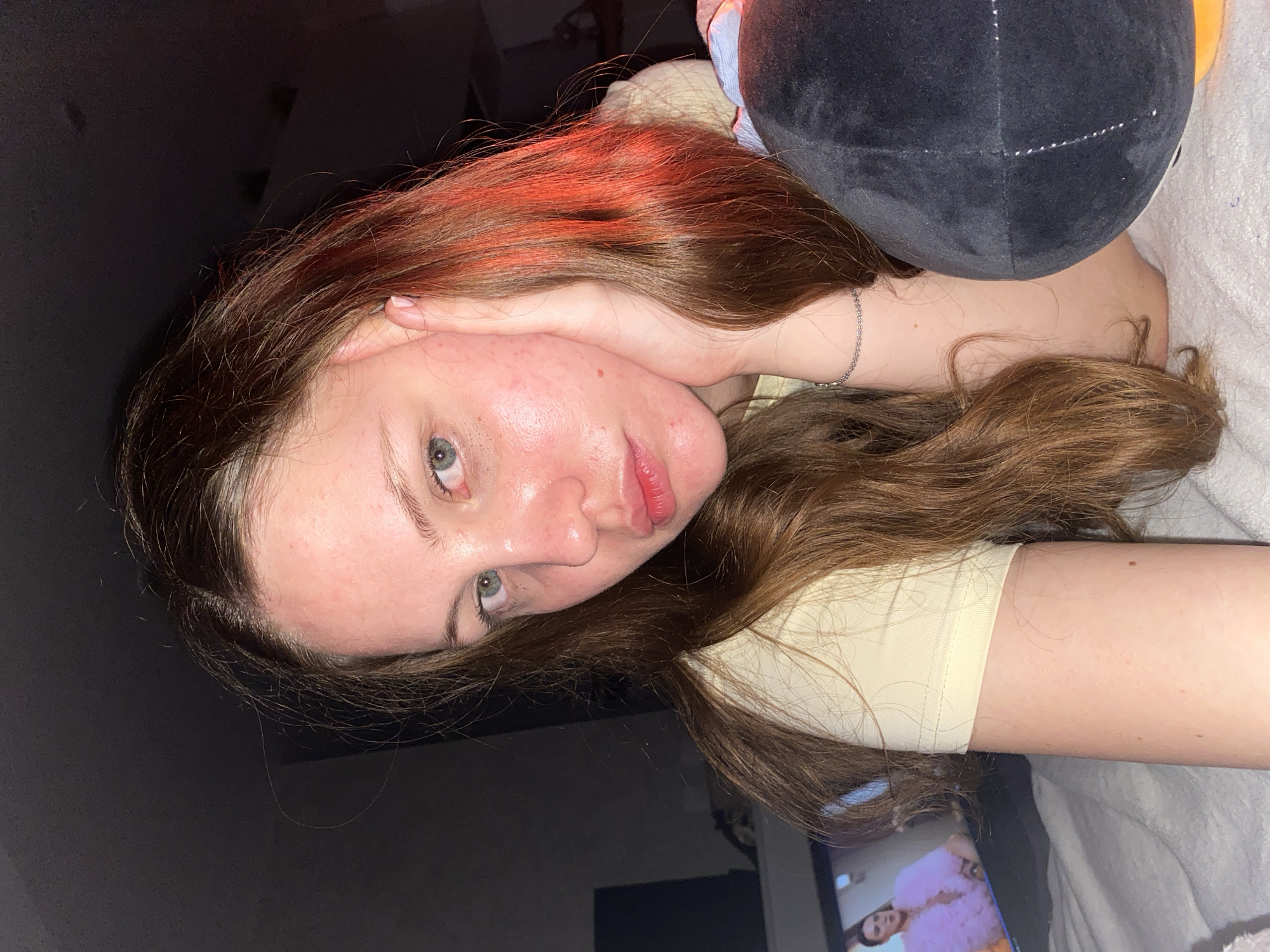 A young woman with brown hair and light green eyes is lying on a bed, resting her head on her hand. She is wearing a light yellow t-shirt. Her face is illuminated by a warm, reddish light, possibly from a lamp. In the background, a television screen displays an image of a person wearing a fluffy pink outfit. A black stuffed toy with white stitching is visible next to her. The environment appears to be a bedroom.