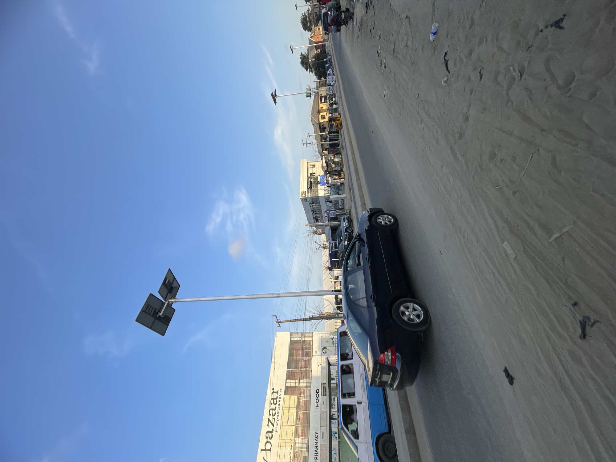 The image shows a street in Ado, Nigeria. The sky is blue with scattered clouds. A black car is parked on the side of the road. A bus is driving near the car. There are buildings and street lights on the side of the road. The road is dirty. There are people in the background and also some cars.