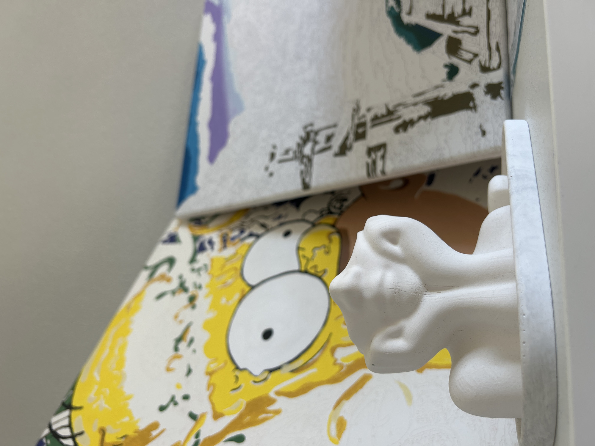 The image shows a white bust of a figure resting on a white shelf. Behind the bust are two paintings or canvases. The left canvas depicts a close-up of Homer Simpson's face, rendered in yellow, white, and black. The right canvas features an abstract landscape with a cloudy sky in shades of blue and purple, and a building-like structure in silhouette. The scene is indoors, likely a home environment, with a plain wall in the background. There are no people visible in the image, and no specific location in Kudrovo, Russia, can be identified.