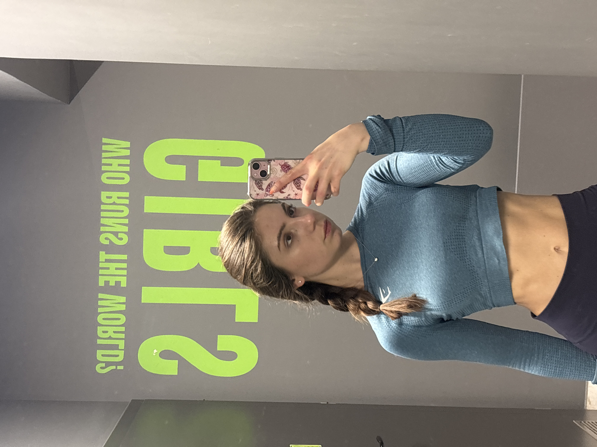 A young woman is captured taking a mirror selfie in what appears to be a fitness studio or gym in Varna, Bulgaria. She is positioned slightly to the left, holding a smartphone with her right hand extended towards the mirror, obscuring part of her face. She looks directly at the phone, with a neutral expression.
She has long, dark brown hair styled in a braid that falls over her right shoulder. She is wearing a form-fitting, teal-blue, long-sleeved crop top, which appears to be athletic wear due to its textured, seamless-knit fabric and a small white logo resembling the Gymshark shark symbol on her chest. The crop top reveals a portion of her midriff and navel. Her lower body is clad in dark-colored leggings or trousers.
Her smartphone has a decorative case featuring light-colored illustrations of sea turtles and other marine-themed patterns. A delicate necklace with a small pendant and stud earrings are visible.
The background is a solid gray wall adorned with large, bright green text, which is reflected in the mirror and therefore appears backwards. When read correctly, the text spells out the phrase: "WHO RUNS THE WORLD? GIRLS." The indoor setting is brightly lit, but the specific time of day is not discernible.