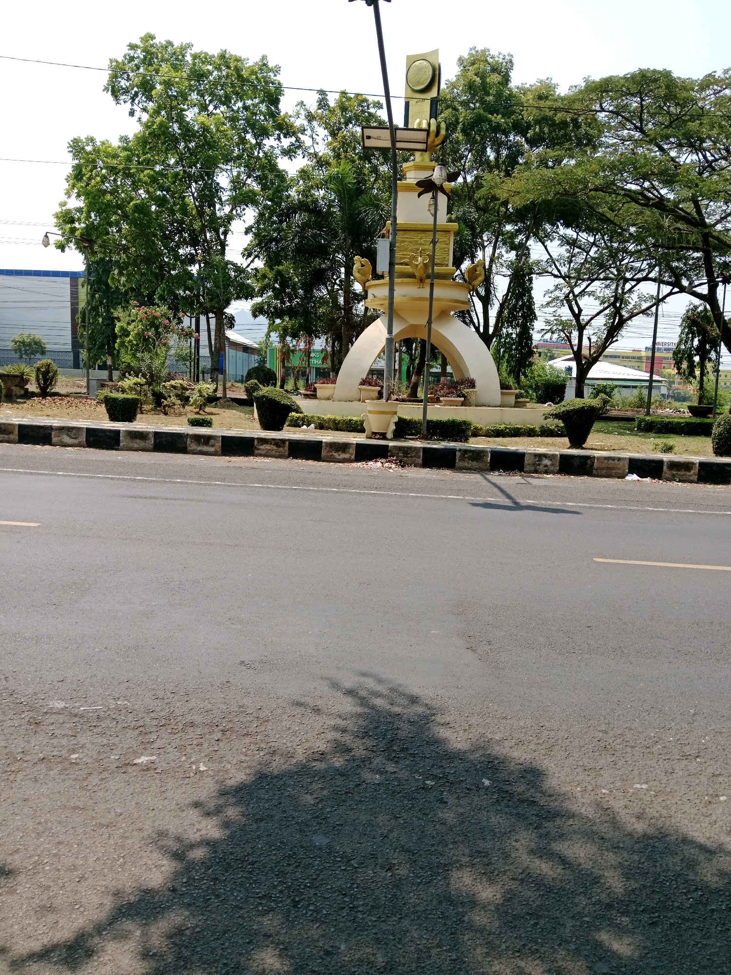 The image shows a monument in the city of Karangtengah, Indonesia, located in front of a building and surrounded by trees. The monument is tall and ornate, with a golden statue on top. There is a paved road in the foreground with a curb and sidewalk. The sky is clear and the sun is shining. The overall scene is one of a well-maintained public space.