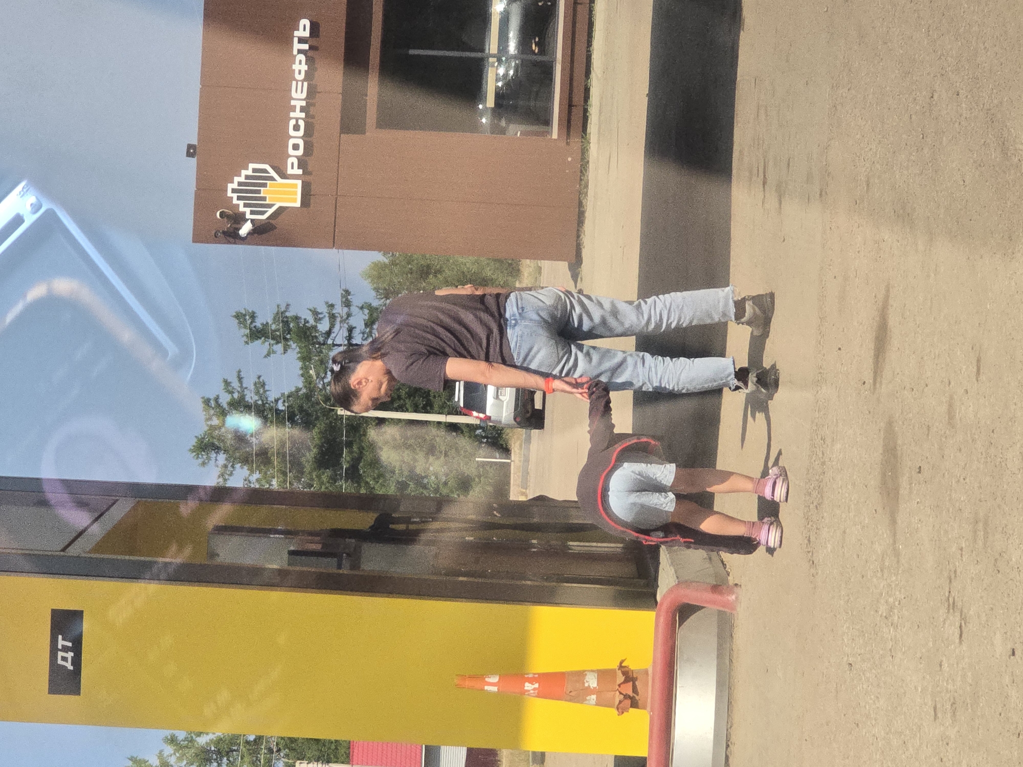 In Turka, Russia, a person with long dark hair tied in a ponytail, wearing a dark t-shirt and light blue jeans, stands near a gas pump. The person is holding the hand of a child who is bent over, facing away from the camera, wearing a gray hoodie and light blue shorts. The child's feet are clad in pink and white striped Crocs. The gas pump is yellow with a black rectangular sign displaying the Cyrillic letters "ДТ". To the right of the gas pump is a brown building with a prominent white logo featuring a stylized flame and the text "РОСНЕФТЬ" in Cyrillic. The ground is paved with asphalt. In the background, trees are visible, and a silver SUV is parked. The scene appears to be outdoors during the daytime, with a clear sky reflected in the glass of the building.