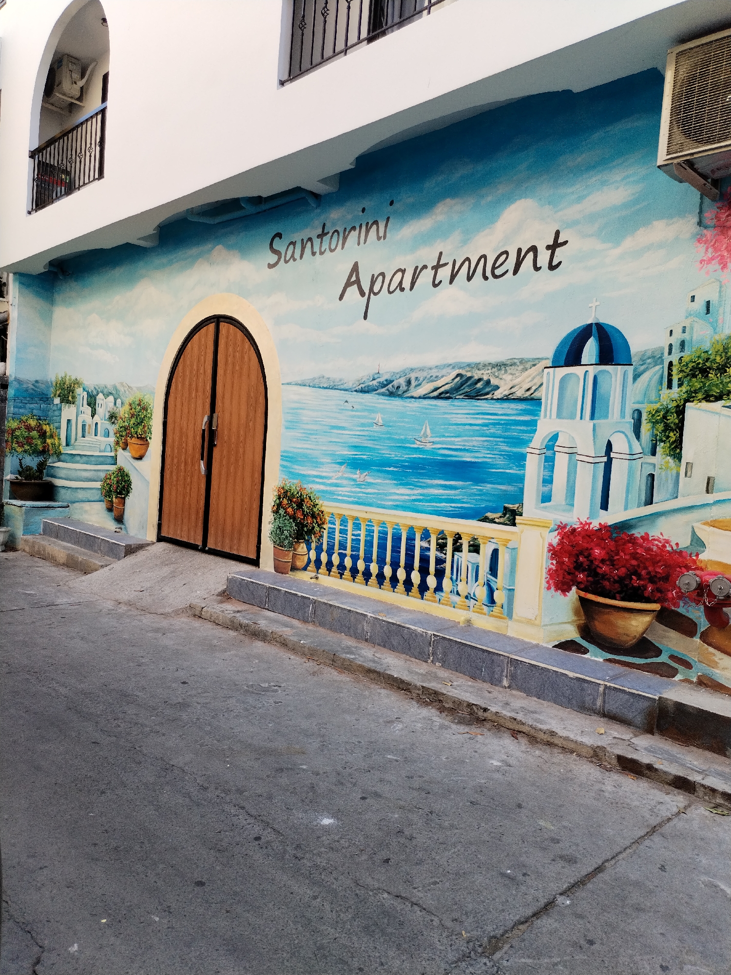 The image shows the exterior of a building in Nha Trang Ward, Vietnam, featuring a painted mural advertising "Santorini Apartment." The mural depicts a Santorini-style scene with white buildings, blue domes, and a seascape with sailboats. A set of brown doors are painted to look like a doorway into the scene. Potted plants are strategically placed to enhance the illusion. The building facade is white, with a small balcony and an air conditioning unit visible above the mural. Below the mural is a paved street and a raised curb. There are no people visible in the image.