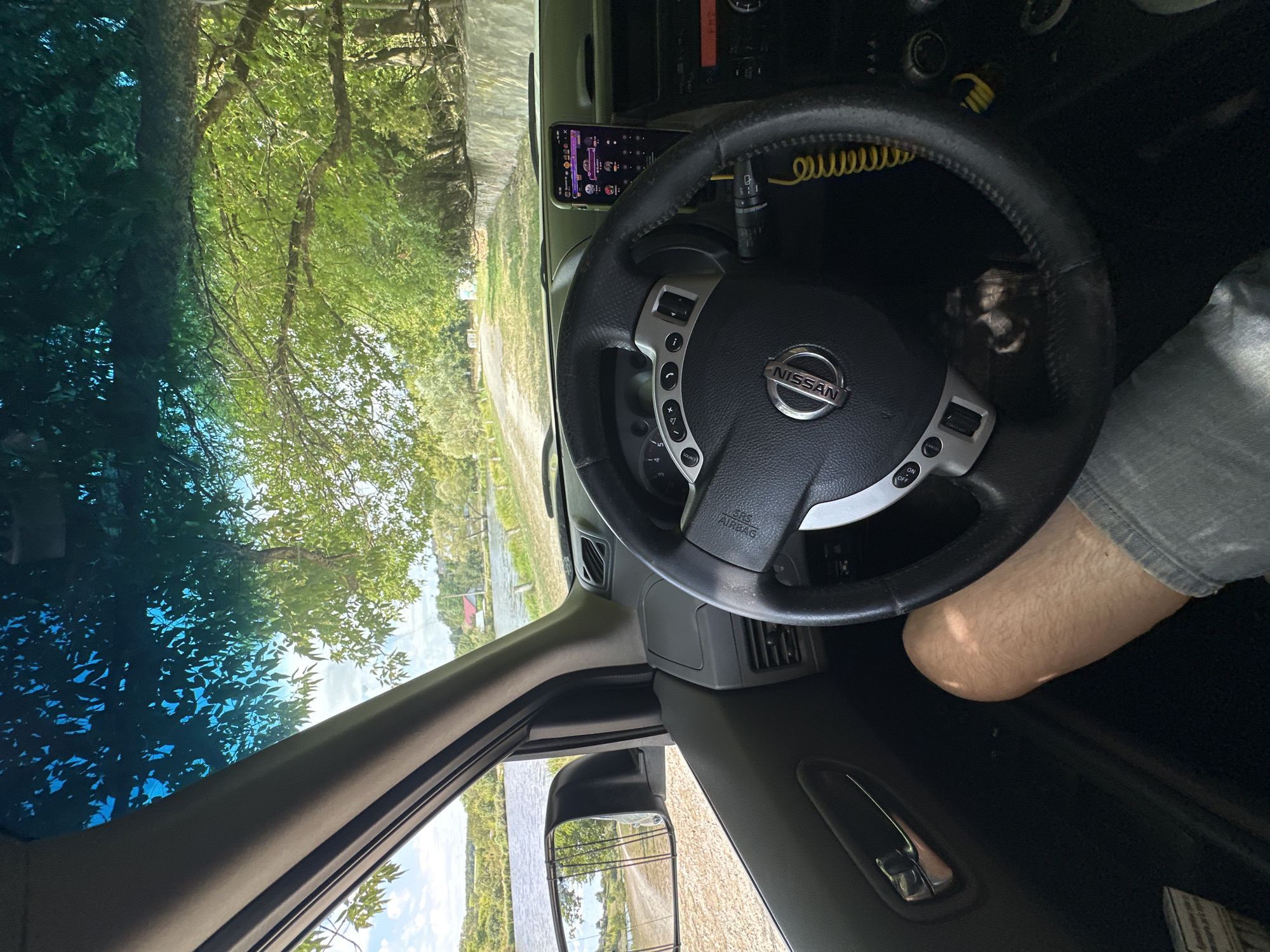 The image shows the interior of a car, likely a Nissan, with the driver's perspective. The steering wheel is prominent in the foreground, with the Nissan logo visible. A smartphone is mounted on the dashboard, displaying a colorful interface. The driver's leg, clad in denim shorts, is visible in the lower part of the frame. Through the windshield, a scenic view of a body of water, possibly a river or lake, is seen. The water is bordered by a dirt road and lush green trees. In the distance, rural houses and rolling hills are discernible under a bright, partly cloudy sky. The overall scene suggests a relaxed drive through a picturesque, possibly rural, setting in Makiv, Ukraine, characterized by natural beauty and a peaceful atmosphere.