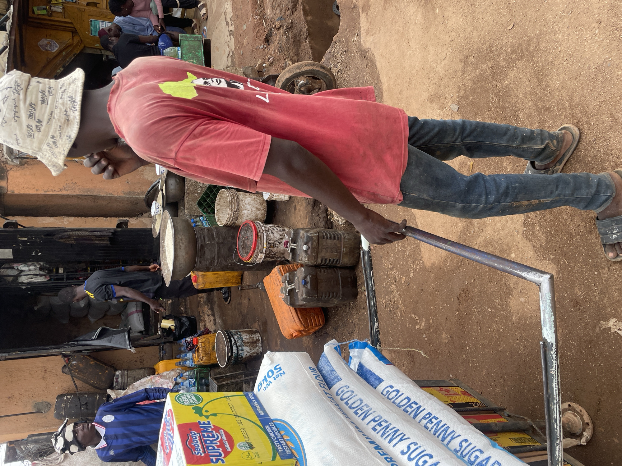 The image depicts a man in Jos, Nigeria, pulling a hand truck laden with goods. He wears a light-colored patterned bucket hat, a red t-shirt, and blue jeans. The hand truck carries bags of "Golden Penny Sugar" and a box of "Supreme Instant Noodles." The surrounding environment is a typical urban scene in Jos, with a dirt ground, assorted containers, and people in the background. Several figures are visible in the background, including one wearing a blue and white jersey.