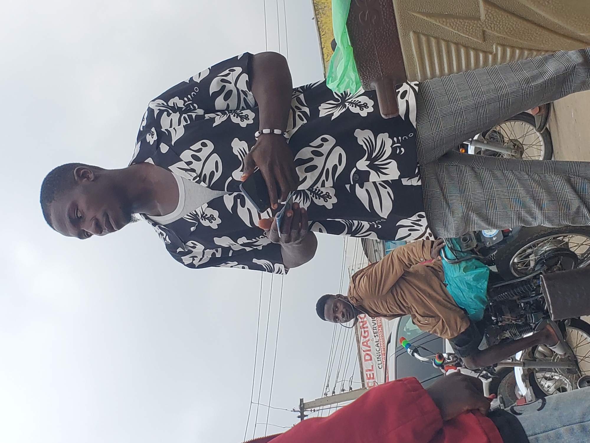 In the city of Gwong, Nigeria, a man wearing a black and white floral shirt and checkered pants stands holding a smartphone. To his left, another man sits on a motorcycle, wearing a brown jacket over black shorts and a turquoise bag on the seat. The background features a cloudy sky, power lines, and a sign for "CEL DIAGNO CLINICAL SERVIC." The scene appears to be a street in a commercial area.
