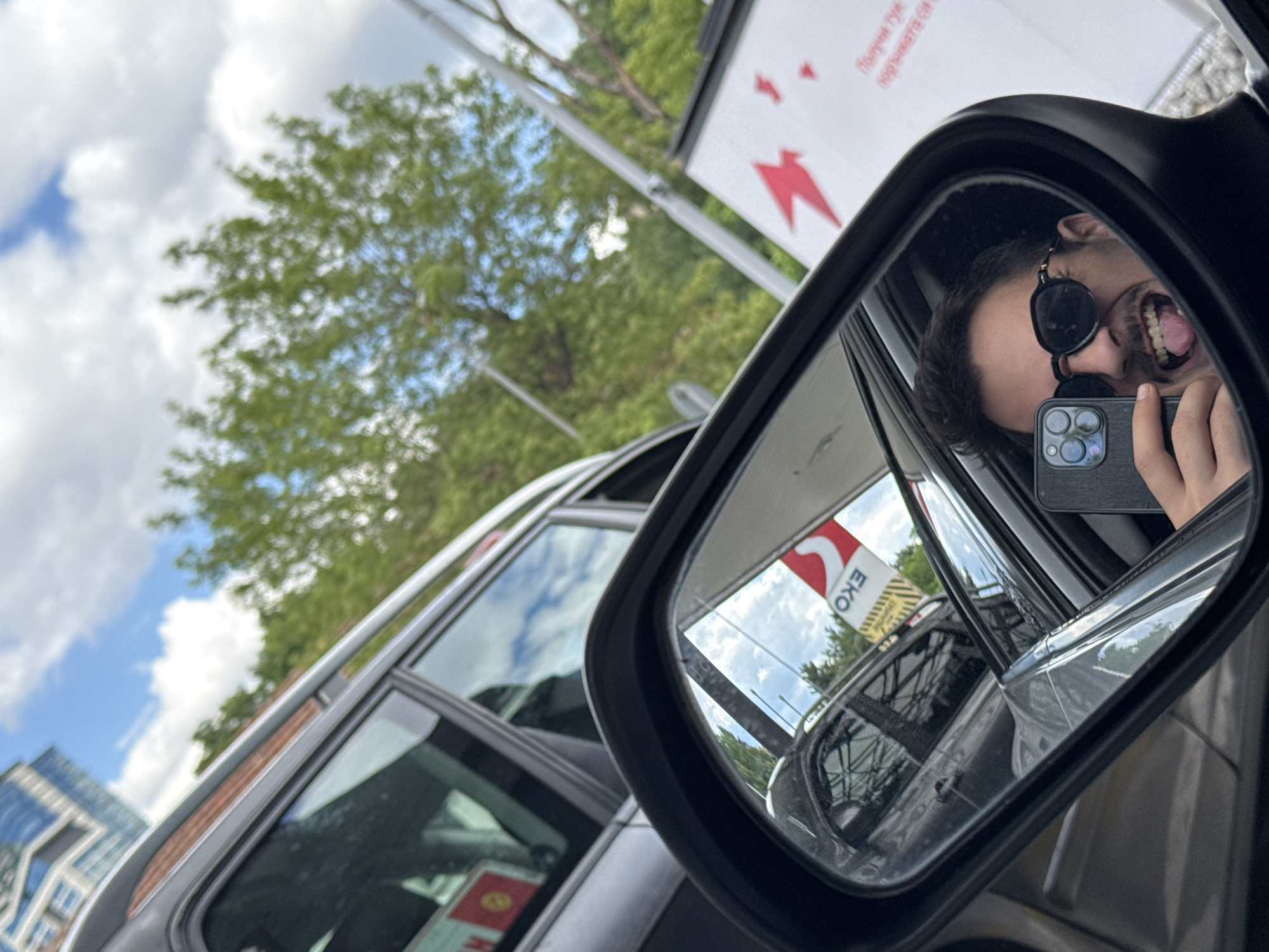 The image captures a selfie taken by a man in a car's side mirror. He is wearing sunglasses and sticking his tongue out. The mirror reflects the man, a smartphone he is holding, and the environment behind him. In the reflection, a red and white sign with the text "EKO" is visible, which is a brand of gas stations. The background outside the car shows trees and a cloudy sky, with a glimpse of a modern building in the distance. This setting is consistent with the outdoor areas of a gas station in Sofia, Bulgaria. The overall scene suggests a casual moment captured by the driver at a service stop.