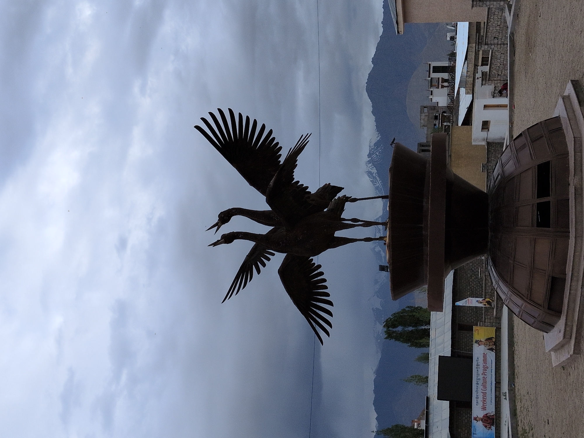 The image depicts a monument of two cranes with outstretched wings, silhouetted against a cloudy sky. The monument is situated in a courtyard or plaza, with buildings and mountainous terrain visible in the background. A banner with the text "Weekend Culture Programme" is displayed on a building to the left of the monument, suggesting the location is likely a cultural center or public space in the city of Leh, India. The overall atmosphere is somber due to the overcast sky and subdued lighting.
