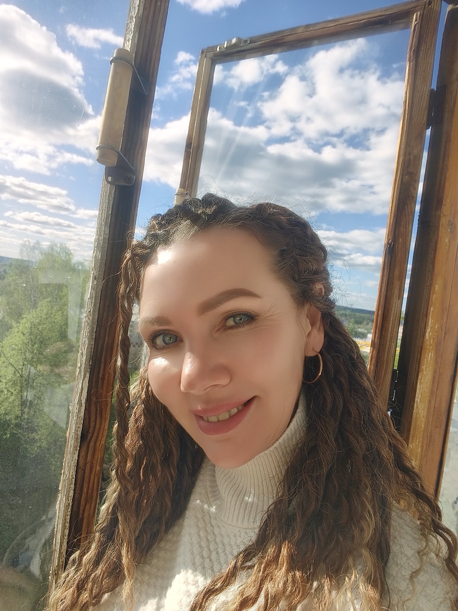 A woman is taking a selfie. She is standing next to a window. The window frame is wooden and partially peeling, indicating it is aged. The window shows a view of a cityscape. The woman has curly hair, braided back at the top, and is wearing a white turtleneck sweater. She is smiling and facing the camera. She has green eyes and gold hoop earrings. The location is Unknown, Unknown.